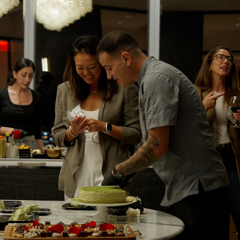 A man with tattoos slices a green cake while a smiling woman watches and holds cards; two other women converse and stand nearby with drinks.