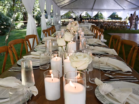 Long wooden table set for an outdoor event with white candles, white roses in glasses, plates, and folded napkins under a white tent.