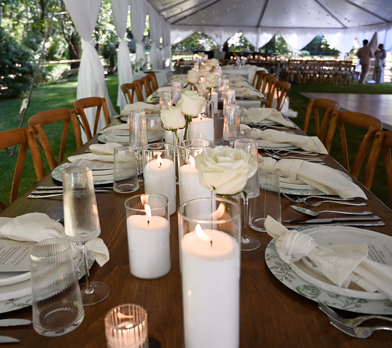 Long wooden table set under a white tent with lit white candles, white roses, and neatly arranged plates, glasses, and cutlery for an outdoor event.