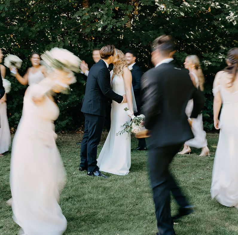 Bride and groom kissing outdoors on grass surrounded by blurred bridesmaids and groomsmen in motion.