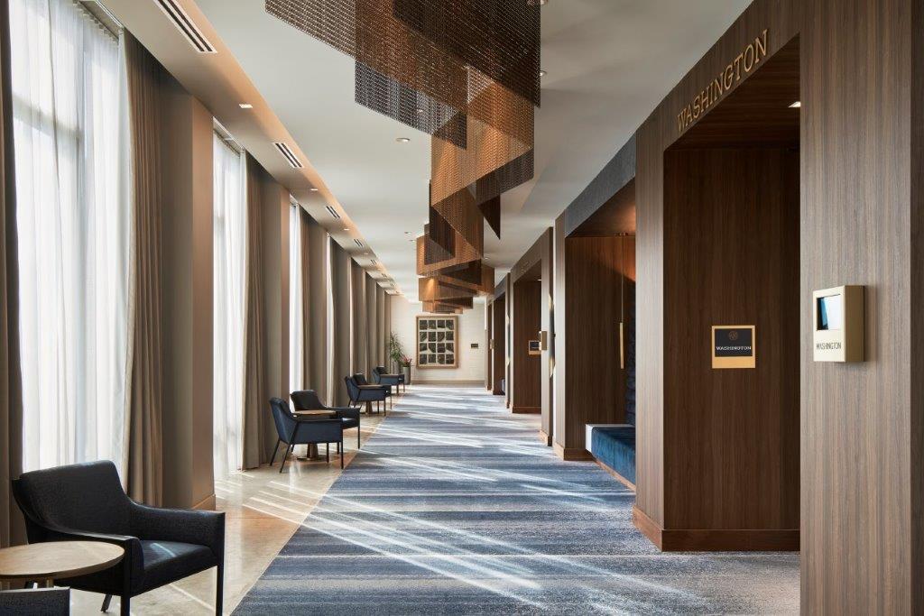 Bright, modern hallway with large windows on the left, blue armchairs along the wall, and wooden alcoves labeled 'WASHINGTON' on the right.
