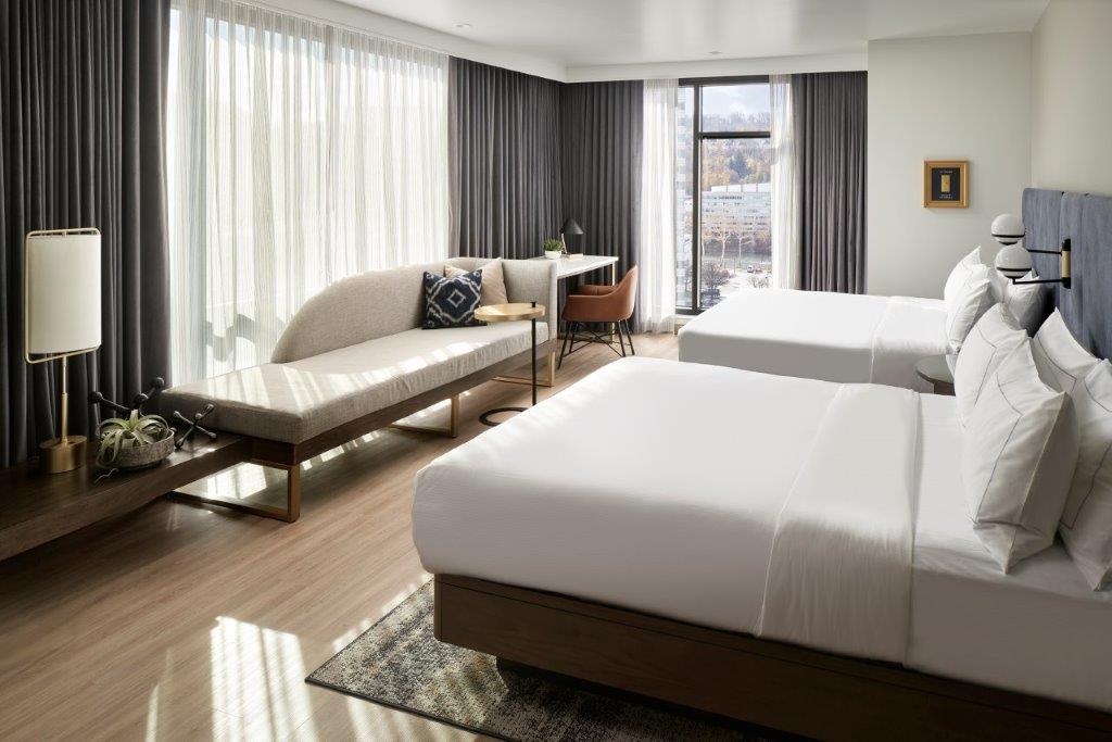 Modern hotel room with two neatly made beds, a curved sofa by large windows with sheer and dark curtains, and a small desk with a chair.