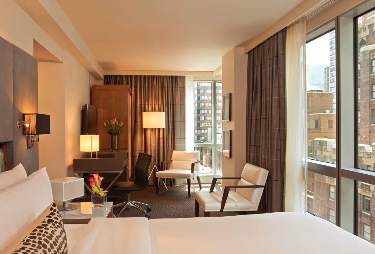 Modern hotel room with white bed, two armchairs, desk with chair, large windows, and city view.