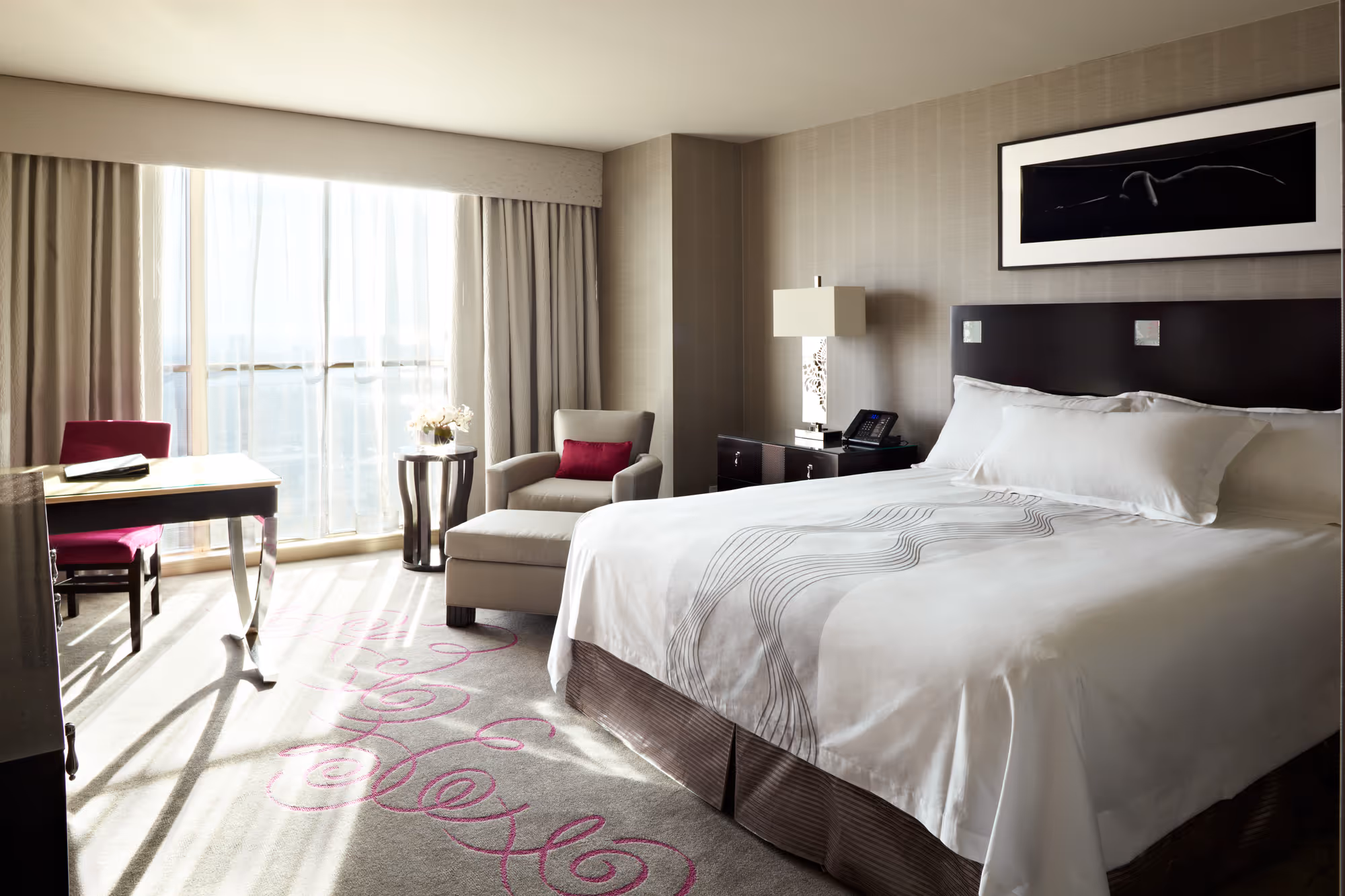 Bright hotel room with a large bed, armchair with a red pillow, desk with red chair, and sheer curtains letting in sunlight.
