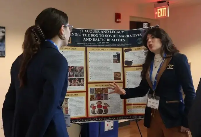 Student presenter explains a research poster on Soviet narratives and Baltic realities to an attendee during a conference session, gesturing while she listens.