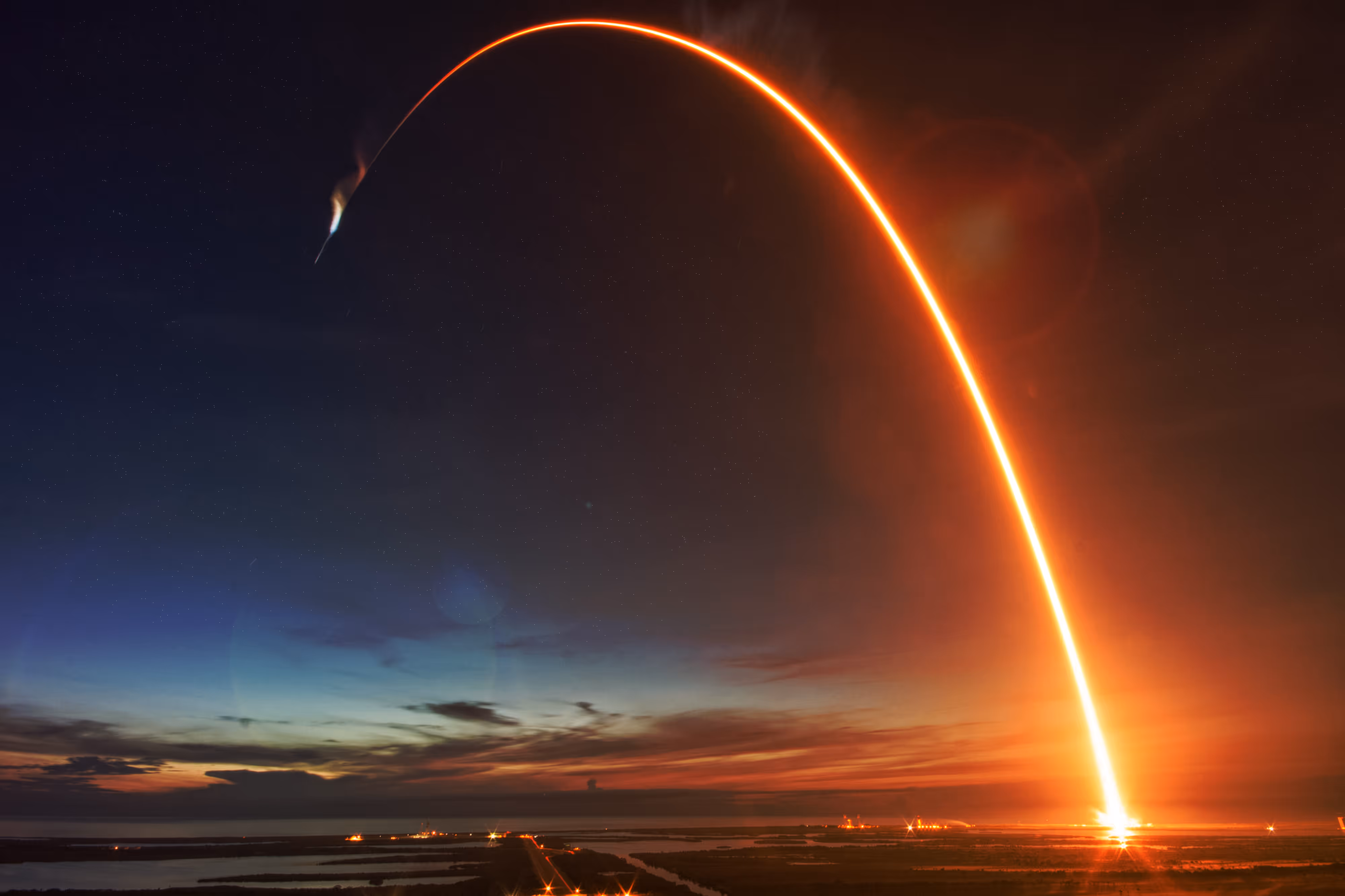 Rocket launching at sunset over the ocean with a bright trail curving upward in the sky.