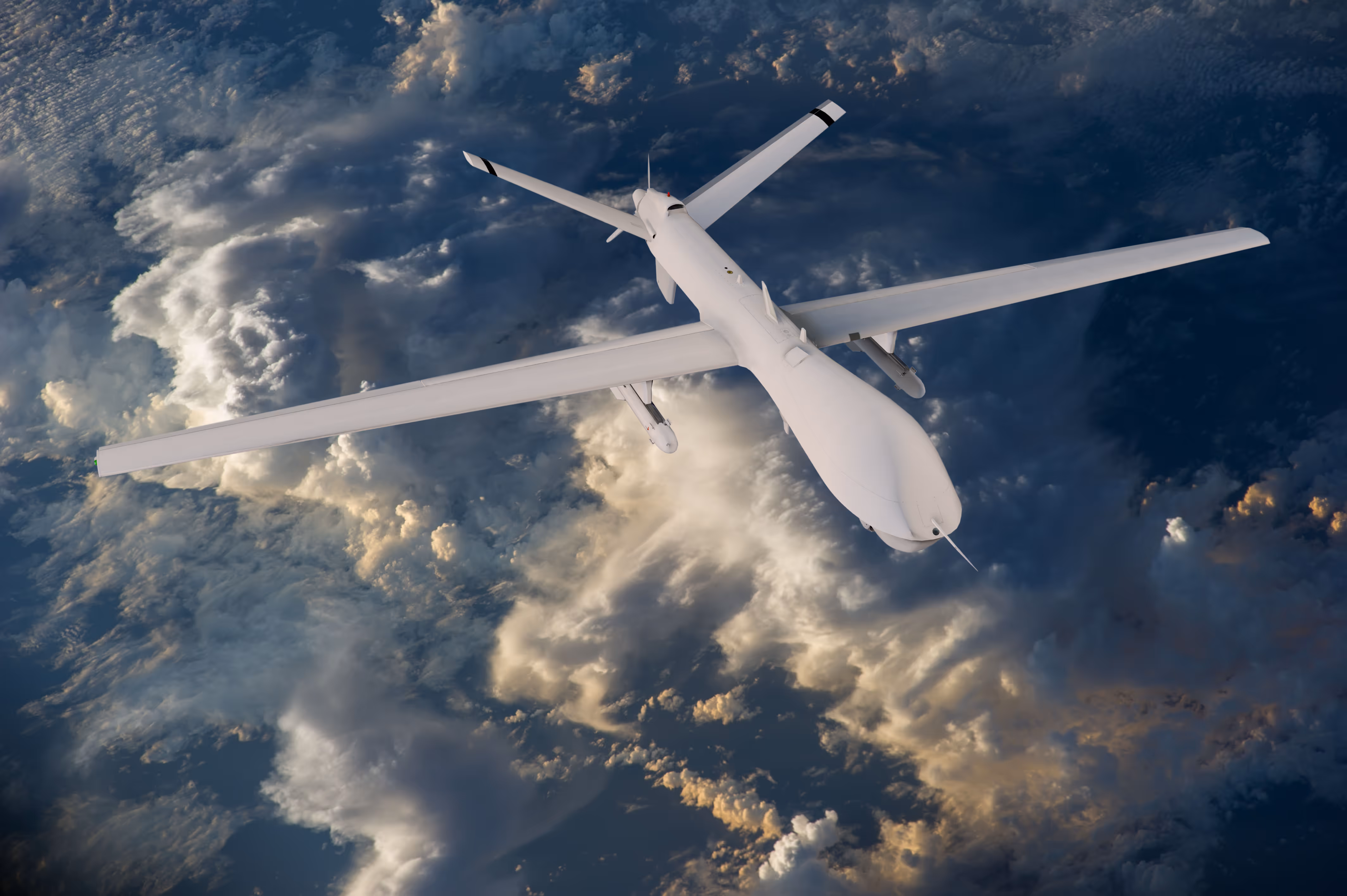 White military drone flying above clouds with sunlight illuminating parts of the aircraft and clouds.