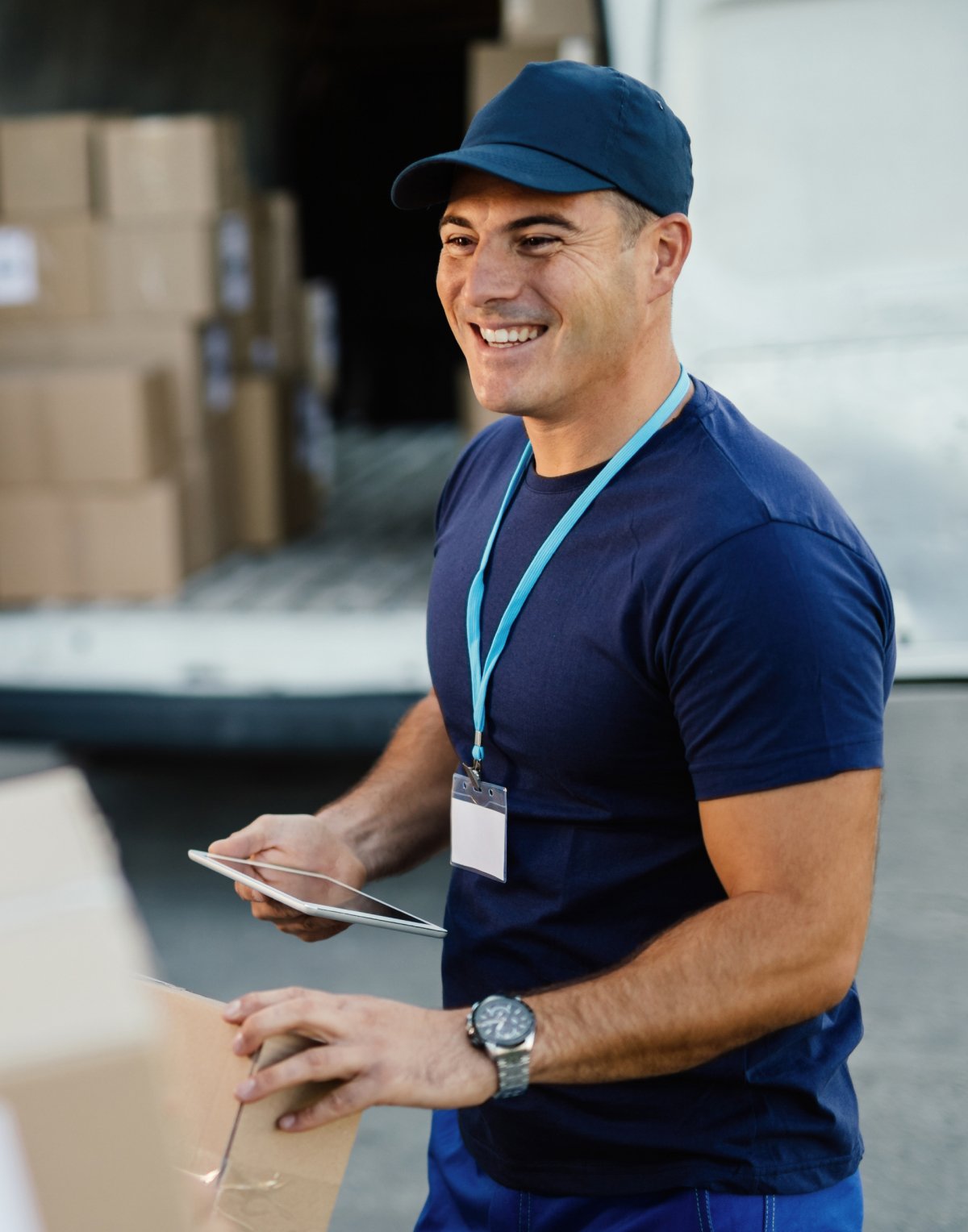 Lächelnder Kurier mit blauer Kappe, der ein Paket hält und ein Tablet benutzt vor einem Lieferwagen mit Kartons.