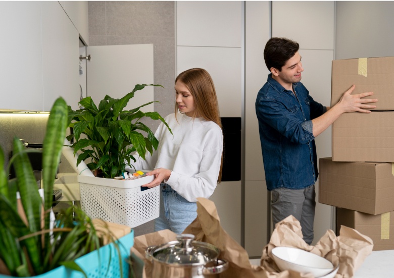 Junges Paar trägt Pflanzen und Kartons beim Umzug in eine Wohnung mit moderner Küche.