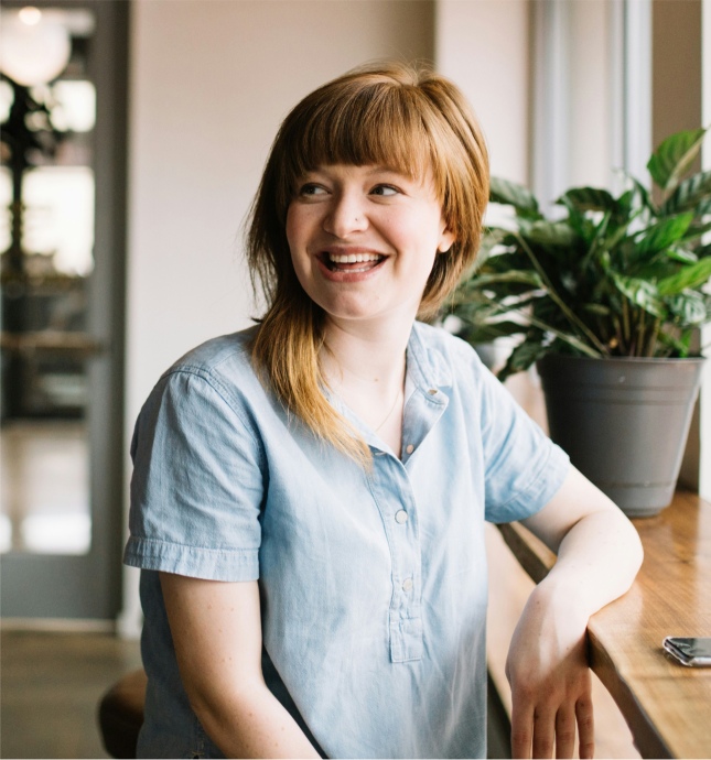 Junge lächelnde Frau mit roten Haaren und hellblauem Hemd sitzt an einem Holztisch neben einer grünen Pflanze.