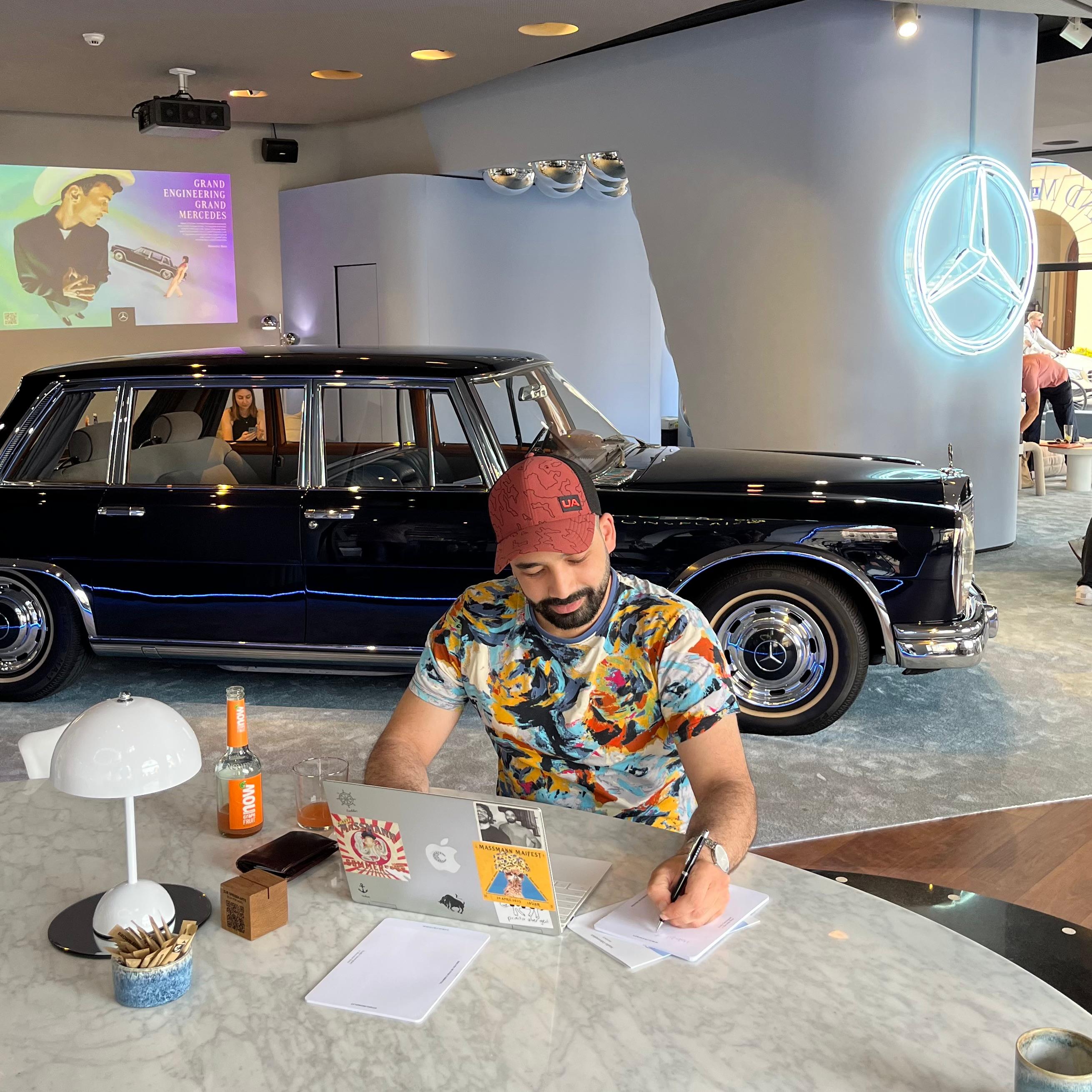 Mann in bunter Shirt und roter Kappe schreibt an einem Marmortisch mit Laptop, vor einem schwarzen klassischen Mercedes in modernem Raum mit Mercedes-Logo-Leuchte.