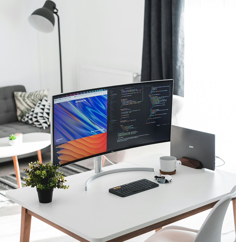 Modern workspace with curved monitor showing code, keyboard, cup, plant, and laptop on white desk.