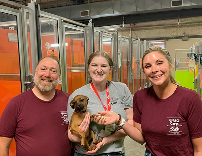 Three smiling people holding a small brown puppy inside an animal shelter.