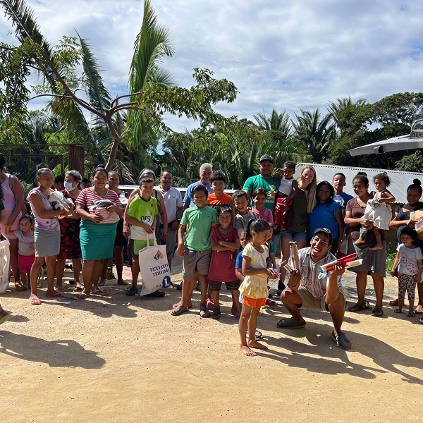 Group of adults and children posing outdoors on sandy ground with tropical trees and cloudy sky.