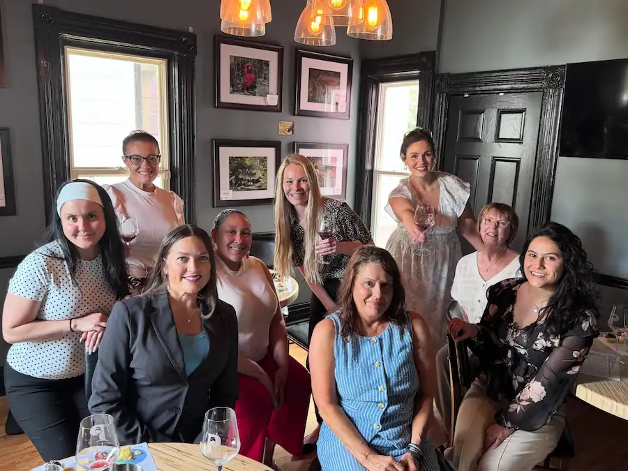 Group of ten women smiling and posing with wine glasses in a cozy room with framed pictures.