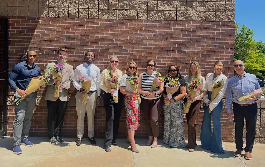 Group of nine people standing outdoors against a brick wall, each holding a bouquet of flowers.