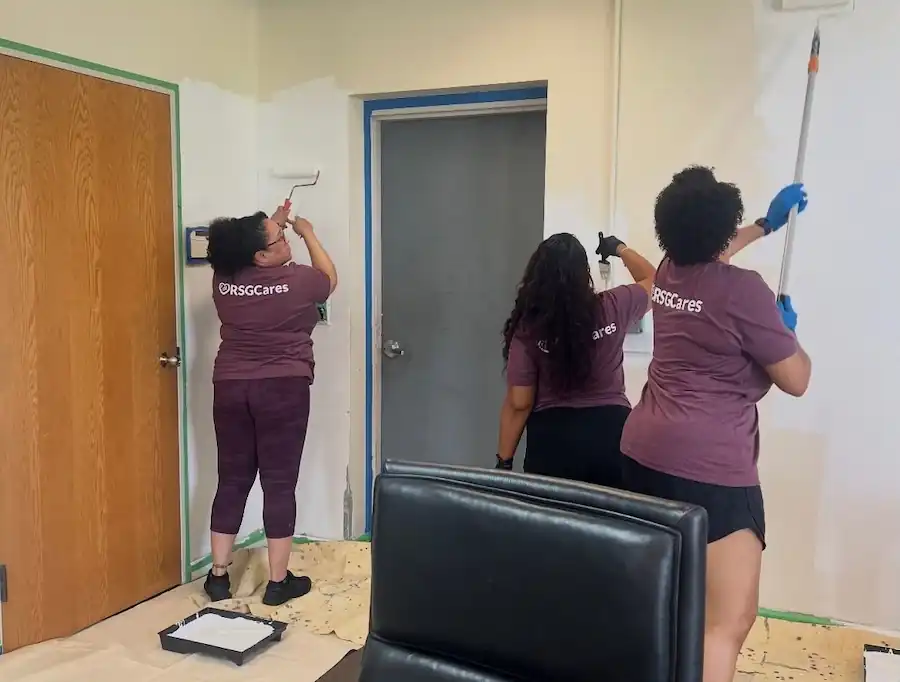 Three women wearing RSGCares shirts painting a white wall in a room with wooden door.