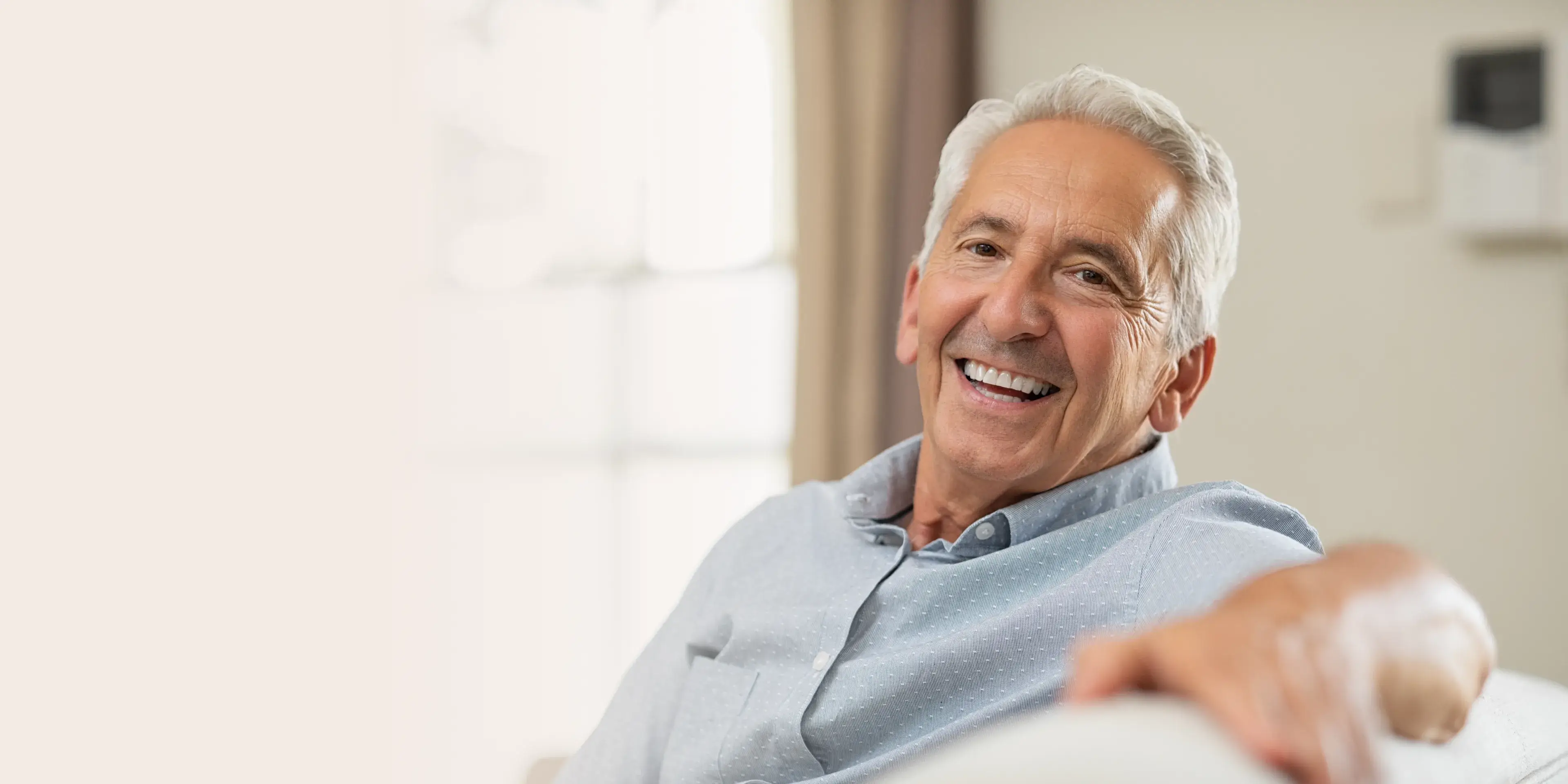 Smiling elderly man with grey hair wearing a light blue shirt, sitting indoors in a bright room.