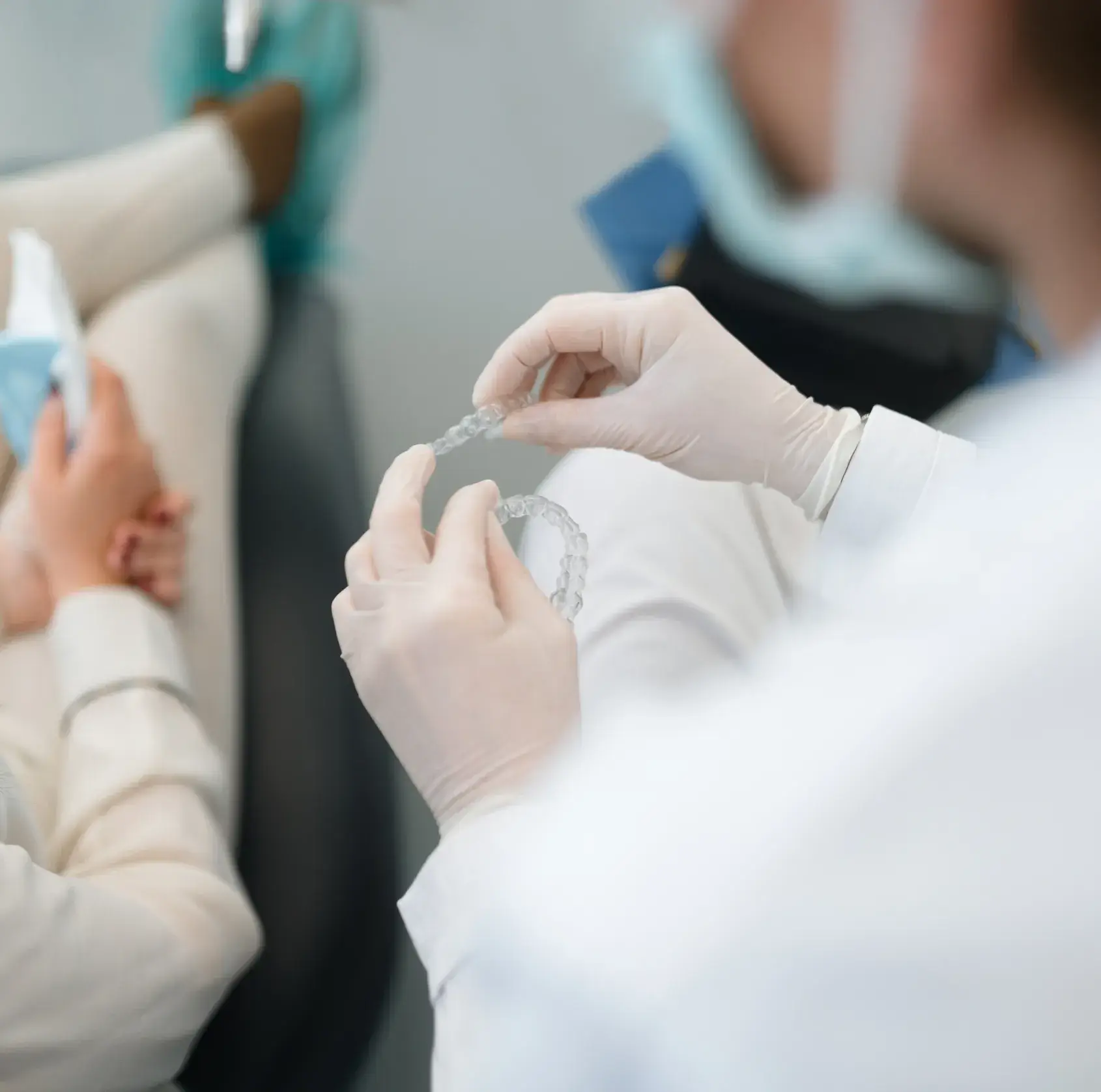 Dentist wearing gloves holding clear dental aligners while a patient sits nearby holding a face mask.