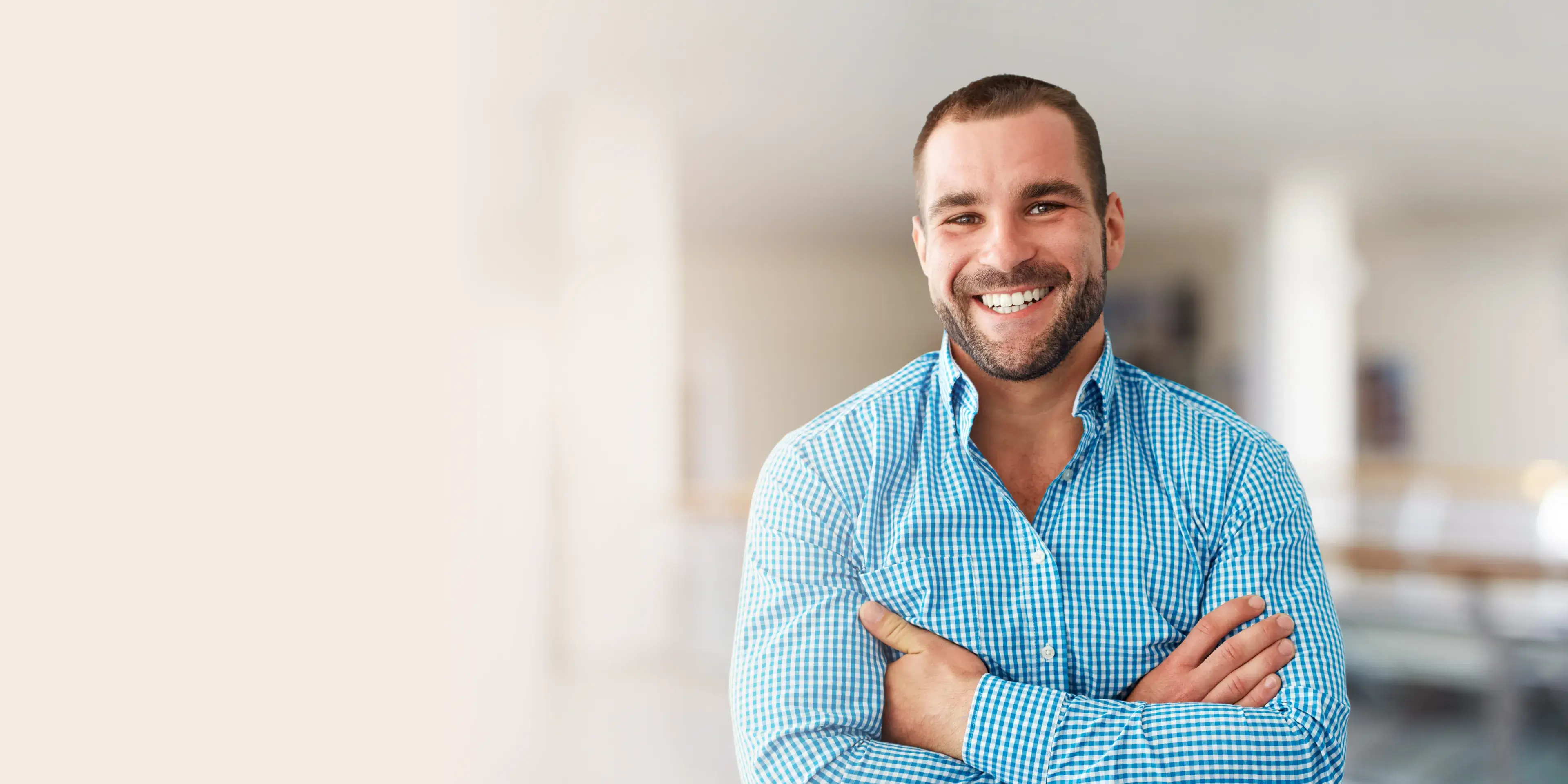 Smiling man in a blue checkered shirt standing with arms crossed in a bright, blurred indoor setting.