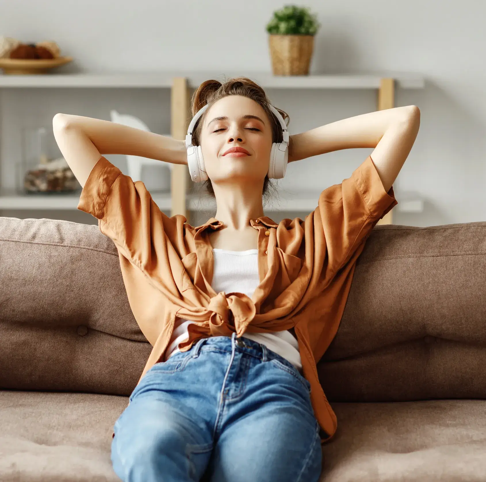 Young woman relaxing with eyes closed wearing headphones on a brown sofa in a cozy living room.