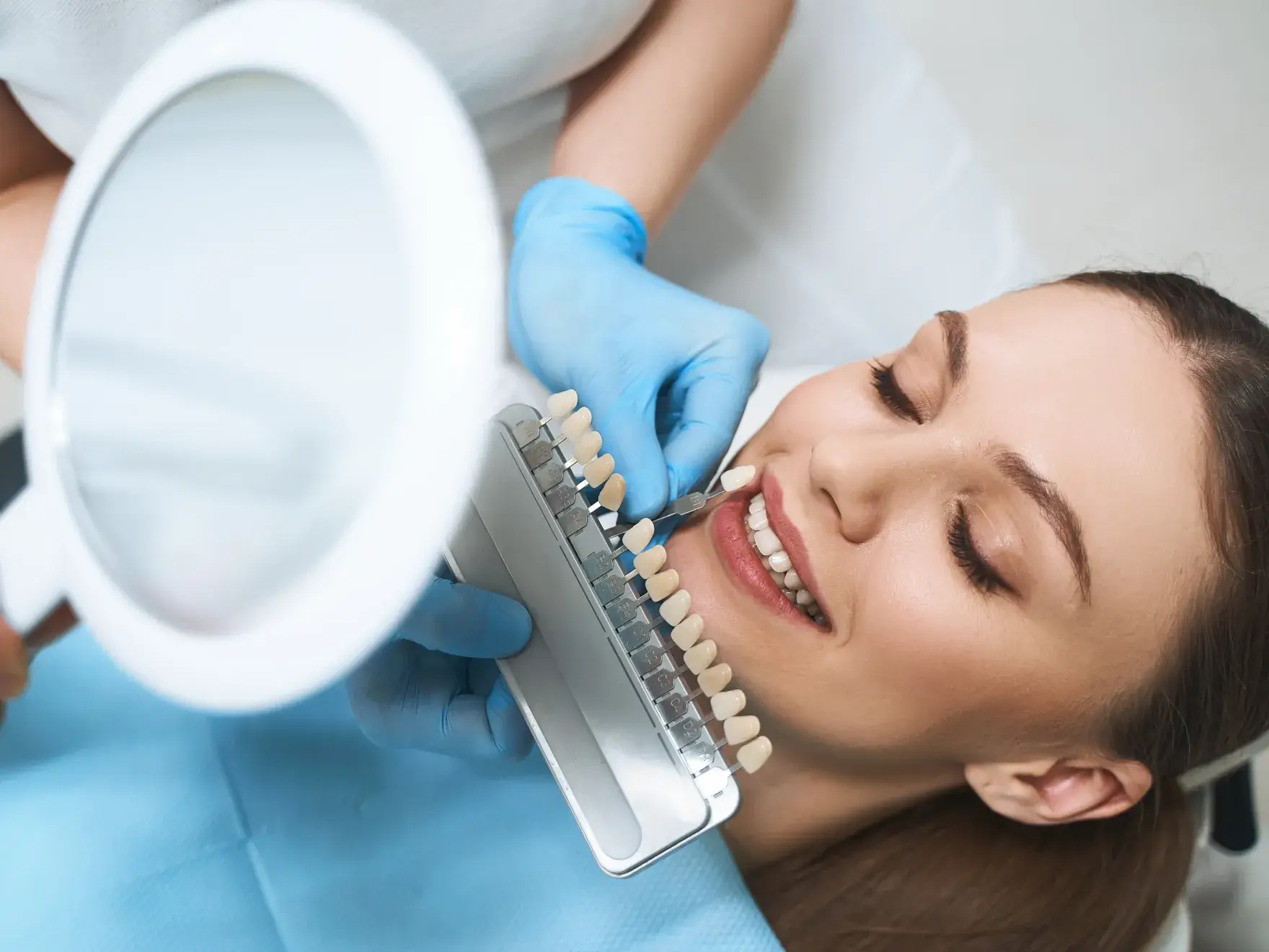 Dentist wearing blue gloves holding a dental shade guide next to a smiling woman's teeth to match veneers.