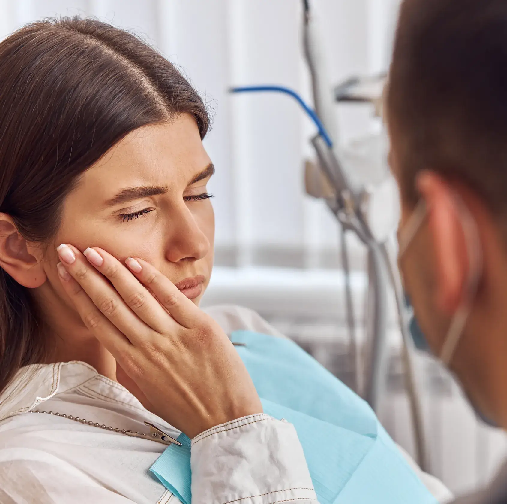 Woman in a dental clinic holding her cheek in pain, with a dentist wearing a mask attending to her.