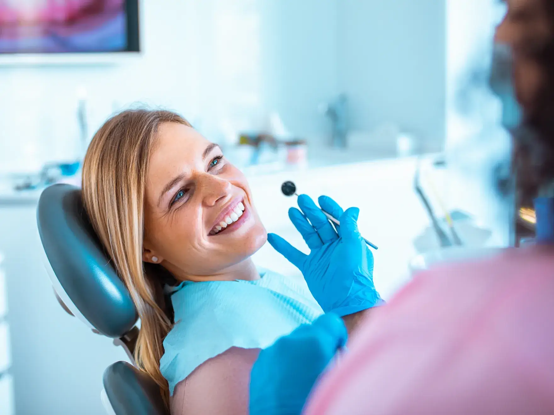 Smiling woman reclining in a dental chair with a dentist holding a dental mirror near her mouth.