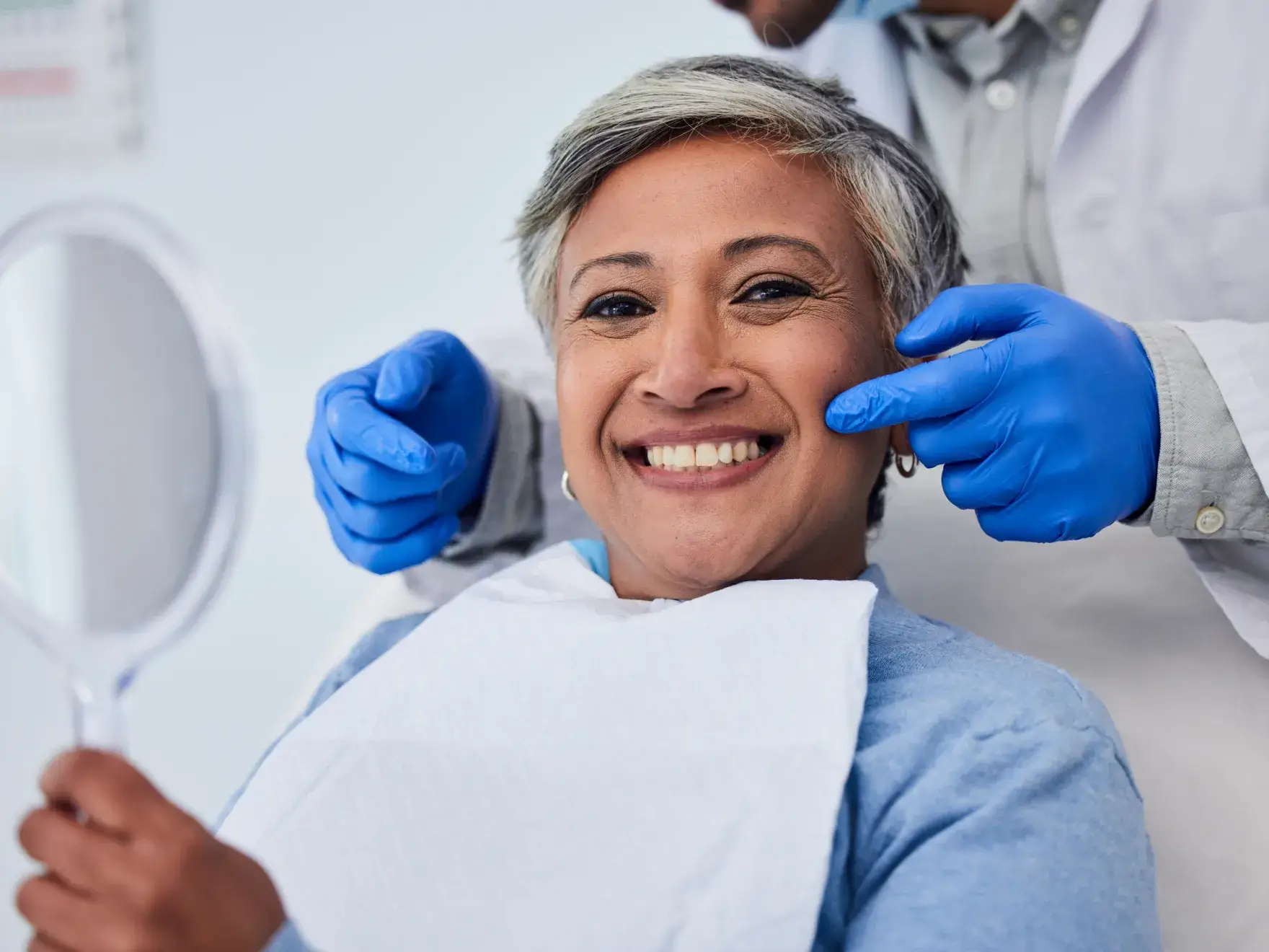 Smiling woman with short gray hair holding a mirror while a dental professional wearing blue gloves touches her cheek.
