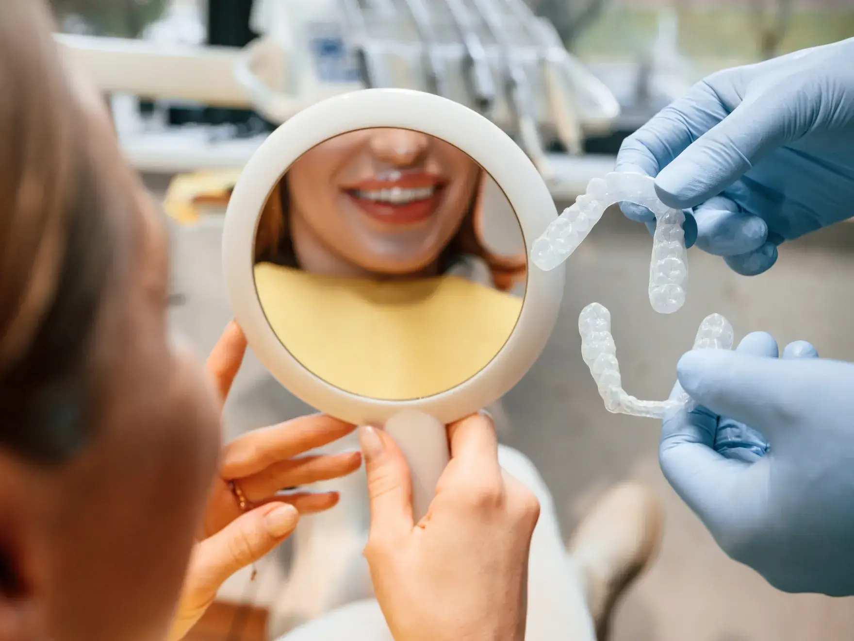 Dentist with blue gloves holding clear dental aligners while patient smiles looking into a round hand mirror.