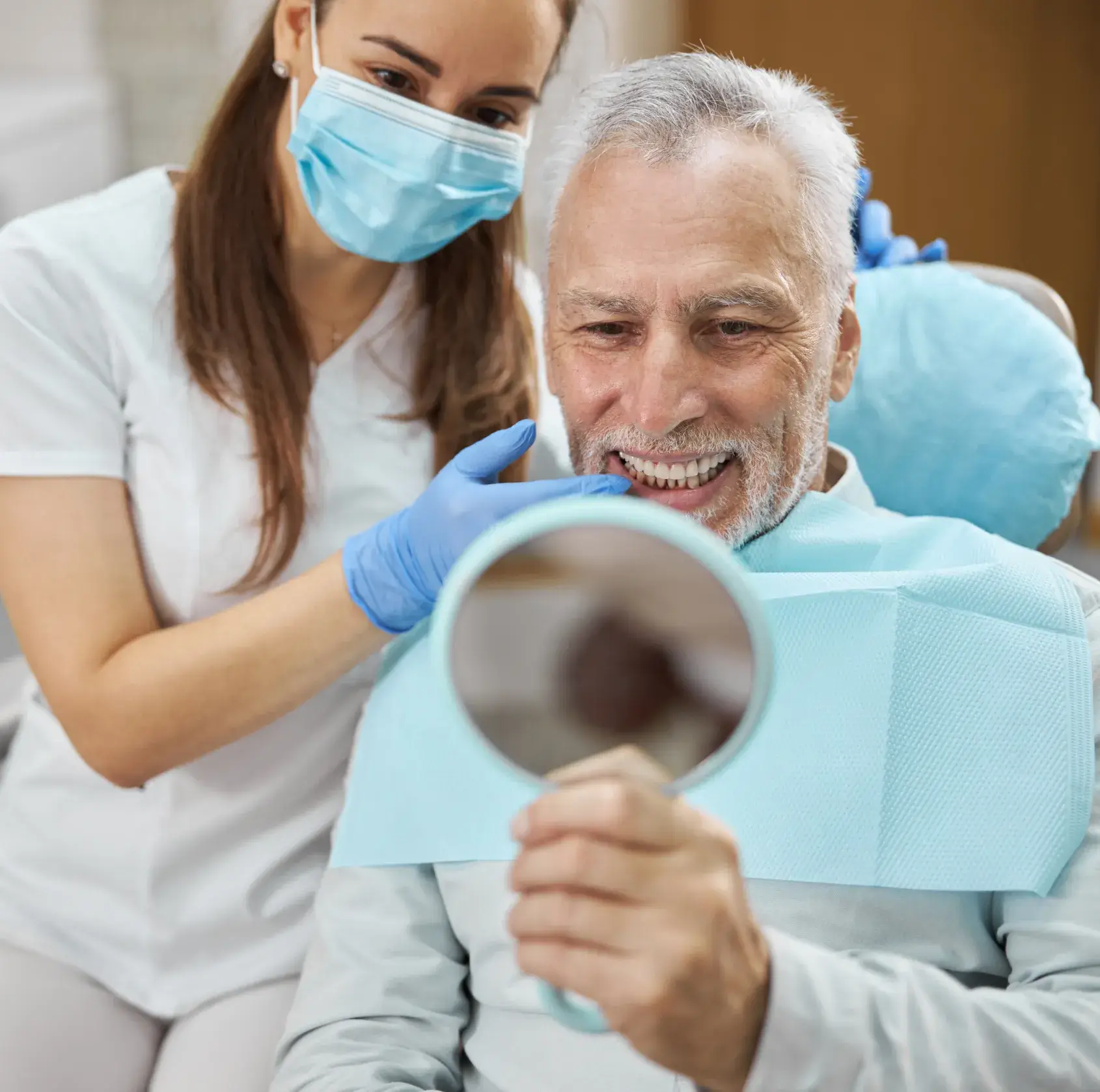 Dentist wearing mask and gloves showing an older man his teeth using a handheld mirror in a dental clinic.