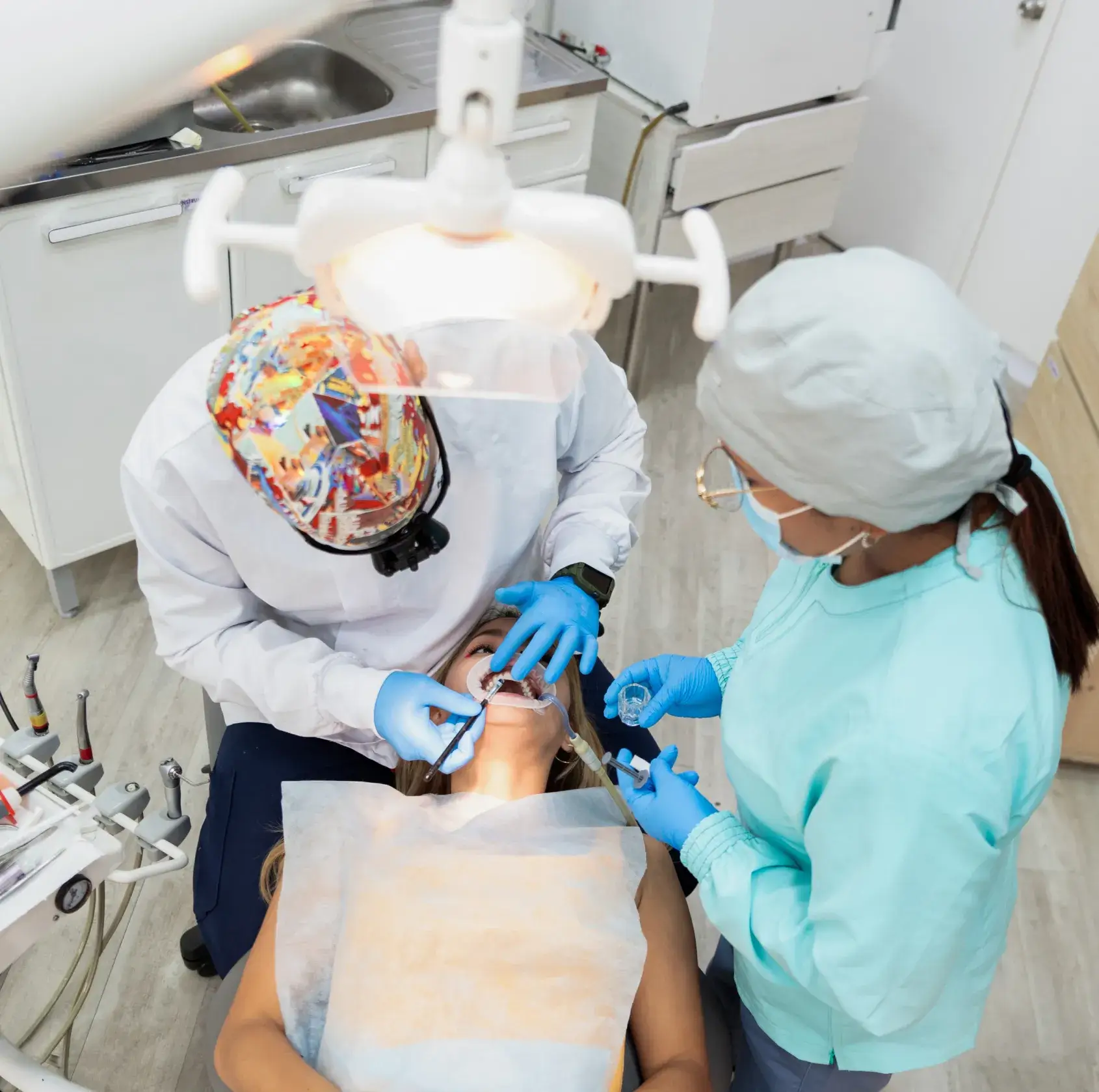 Dentist and assistant wearing protective gear treating a female patient lying in a dental chair under a dental light.