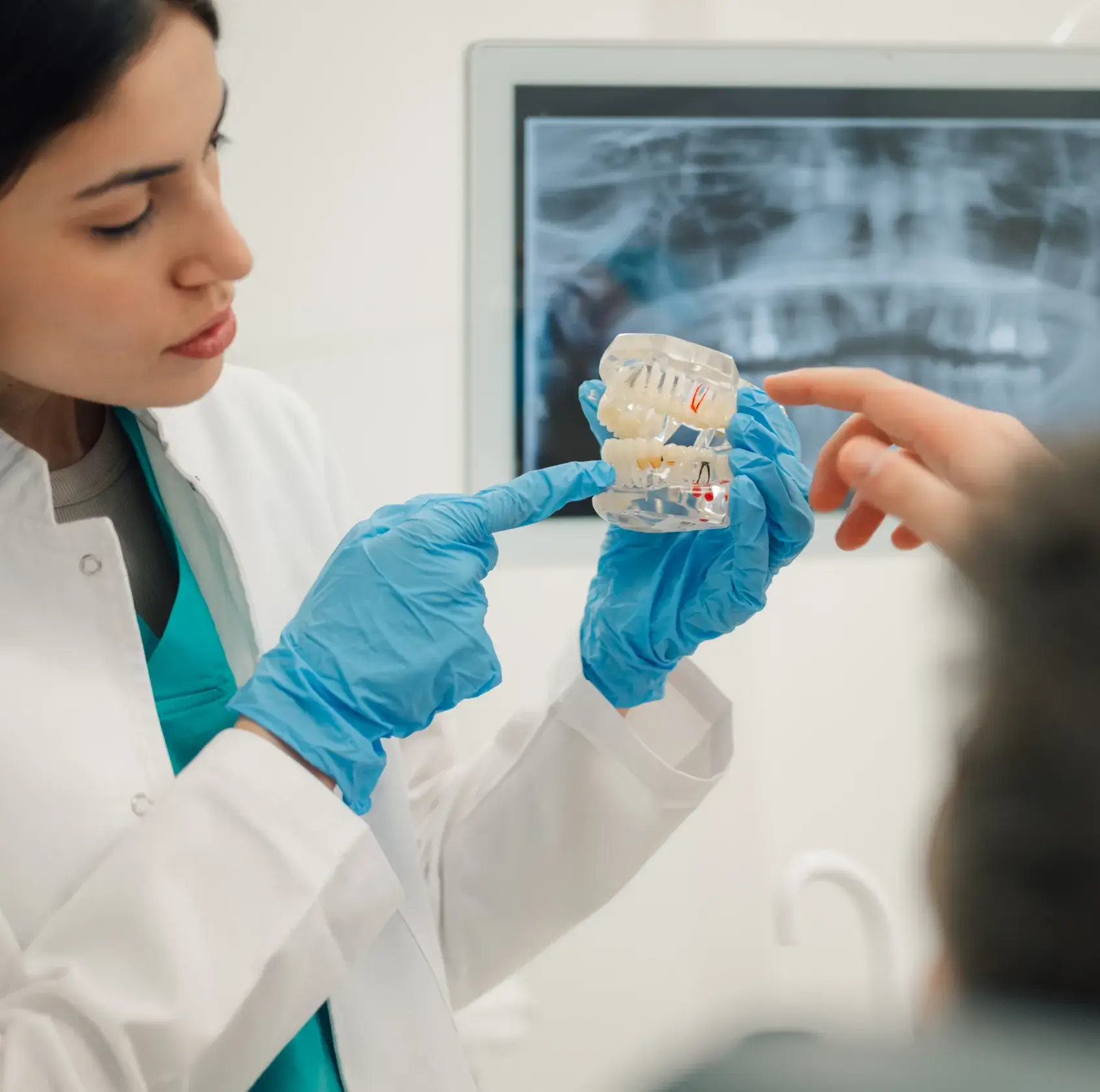 Dentist wearing blue gloves holding a dental model and explaining to a patient with a dental X-ray displayed in the background.