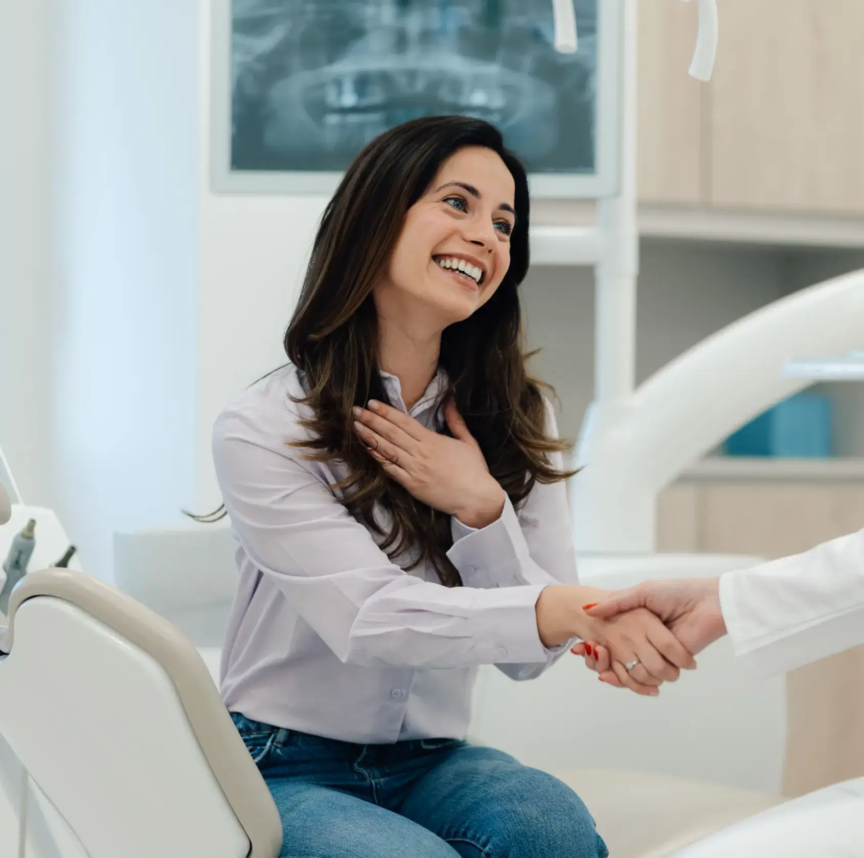 Smiling woman sitting in a dental chair shaking hands with a dentist in a clinical setting.