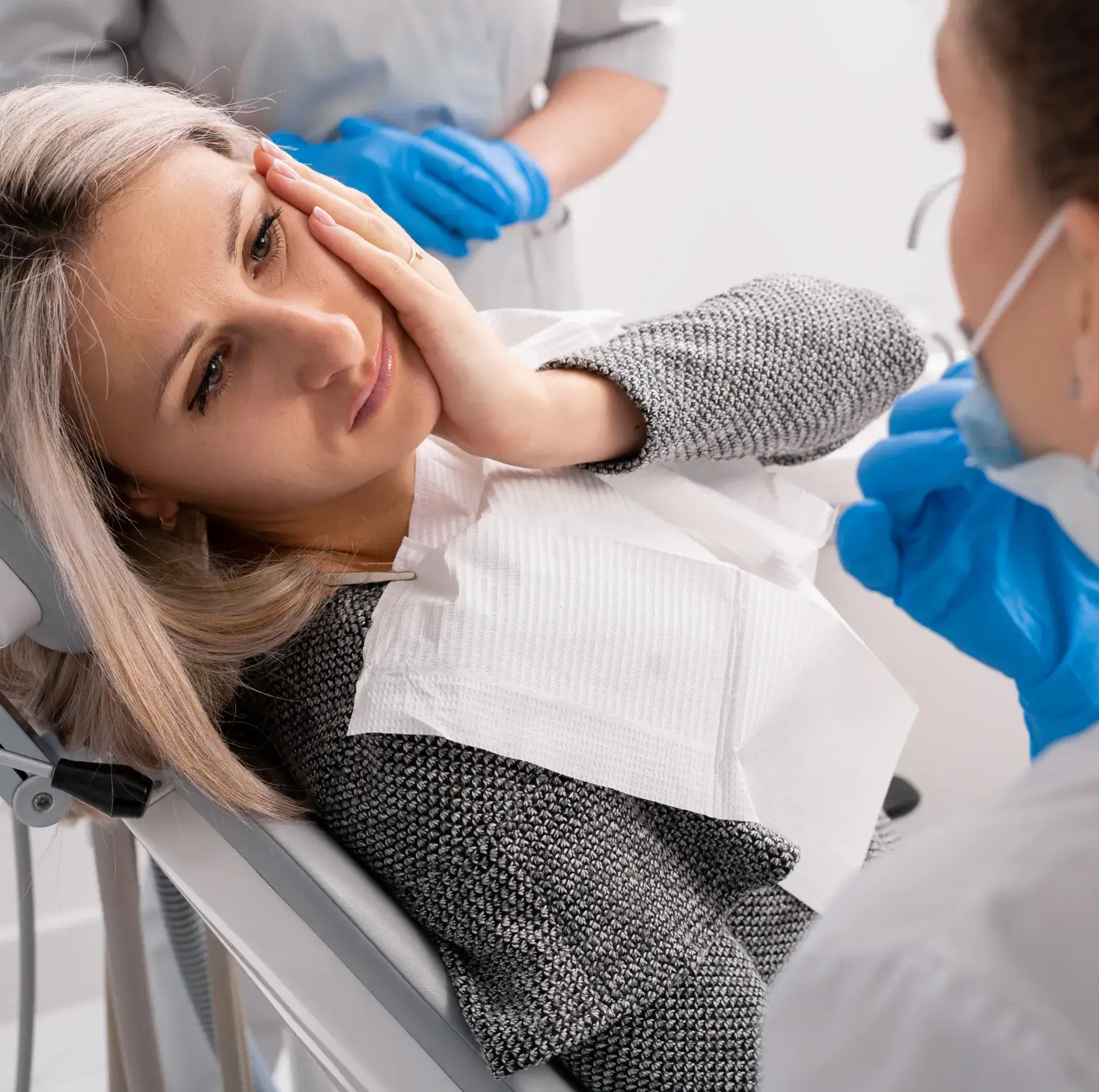 Woman wearing a dental bib sitting in a dental chair, holding her cheek in pain while a dentist wearing blue gloves speaks to her.