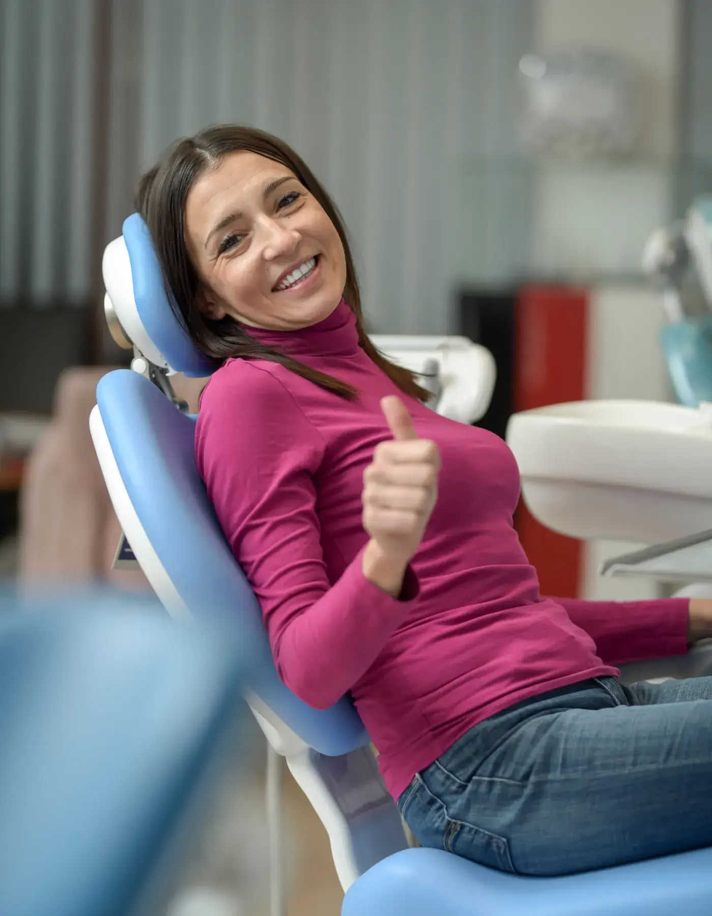 Smiling woman giving thumbs up while sitting in a dental chair in a clinic.