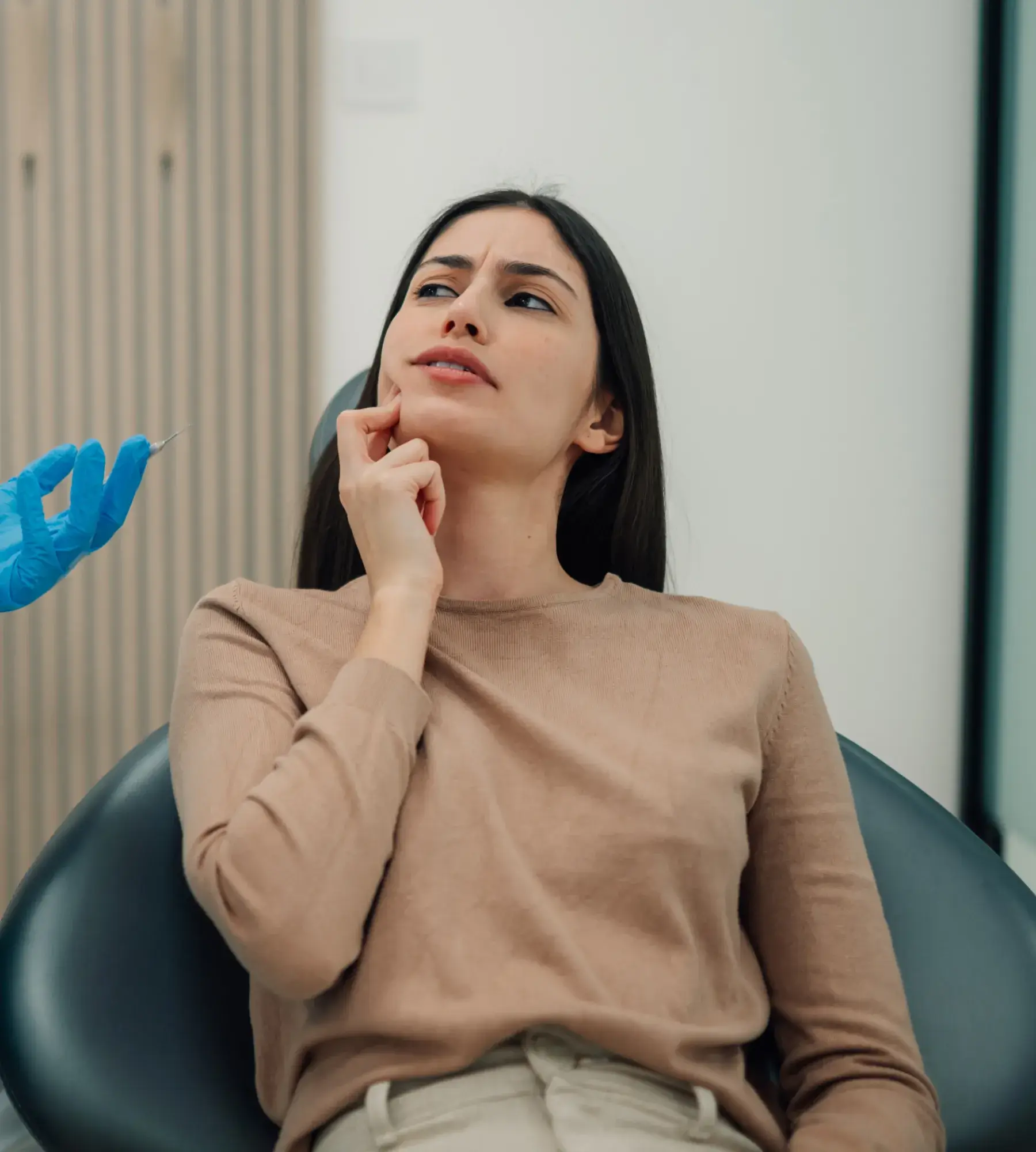 Young woman sits in a dental chair touching her cheek with a concerned expression as a gloved hand holds a dental syringe nearby.
