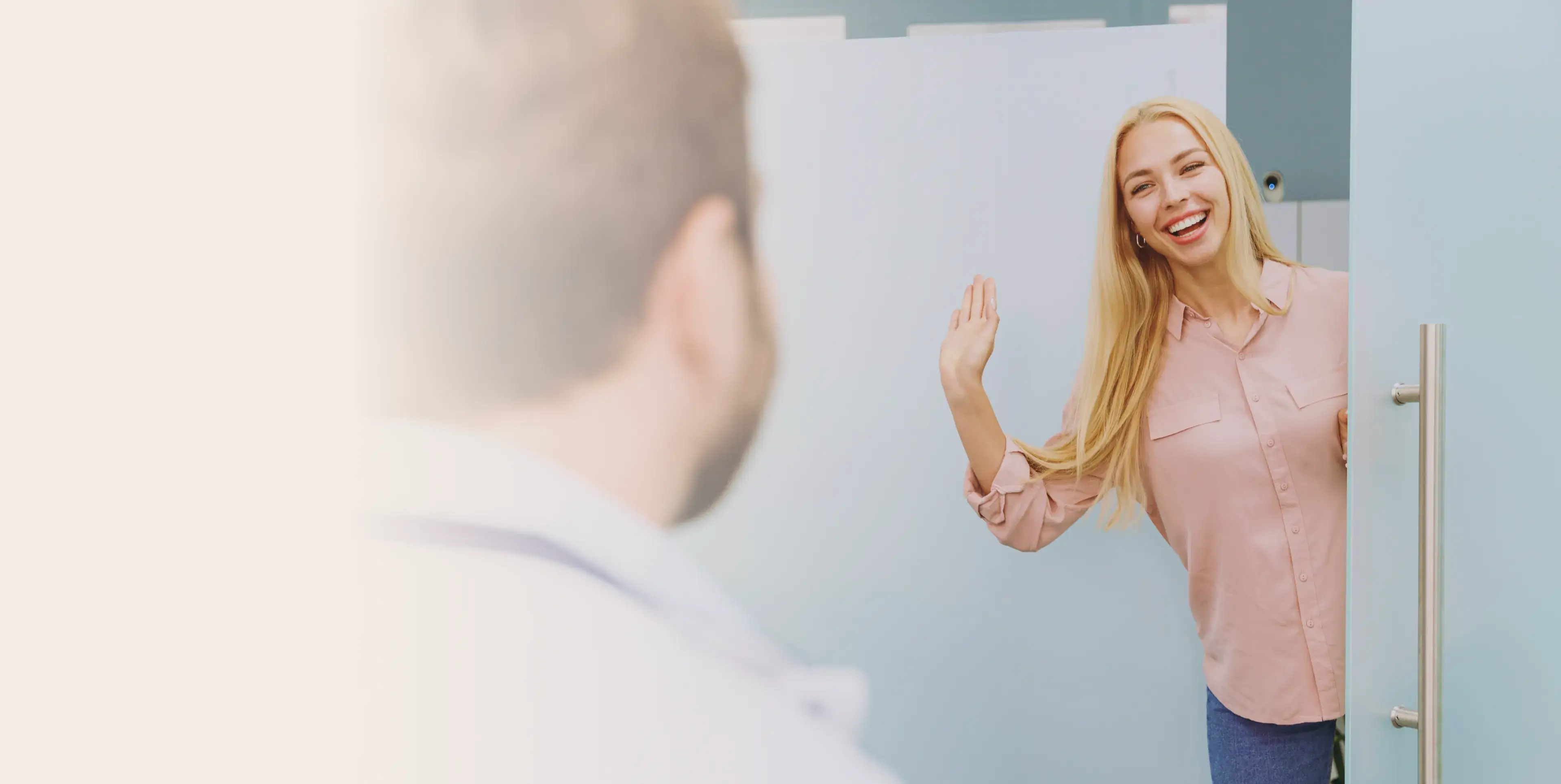 Smiling blonde woman waving while standing in a doorway, facing a man seen from behind.