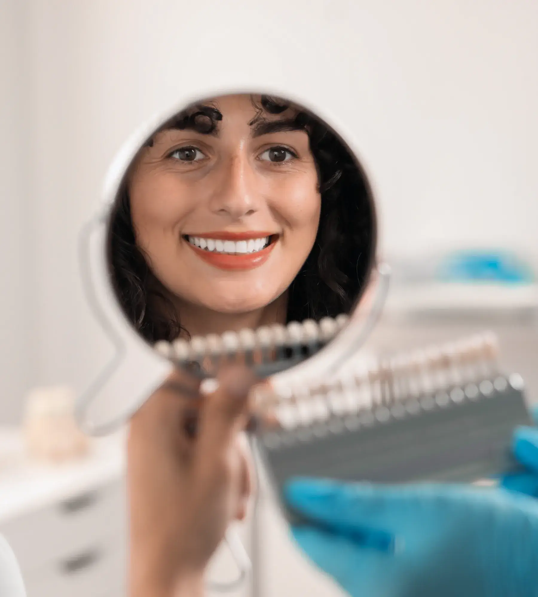 Smiling woman with curly hair looking at her teeth in a round hand mirror held by a gloved hand showing a dental shade guide.