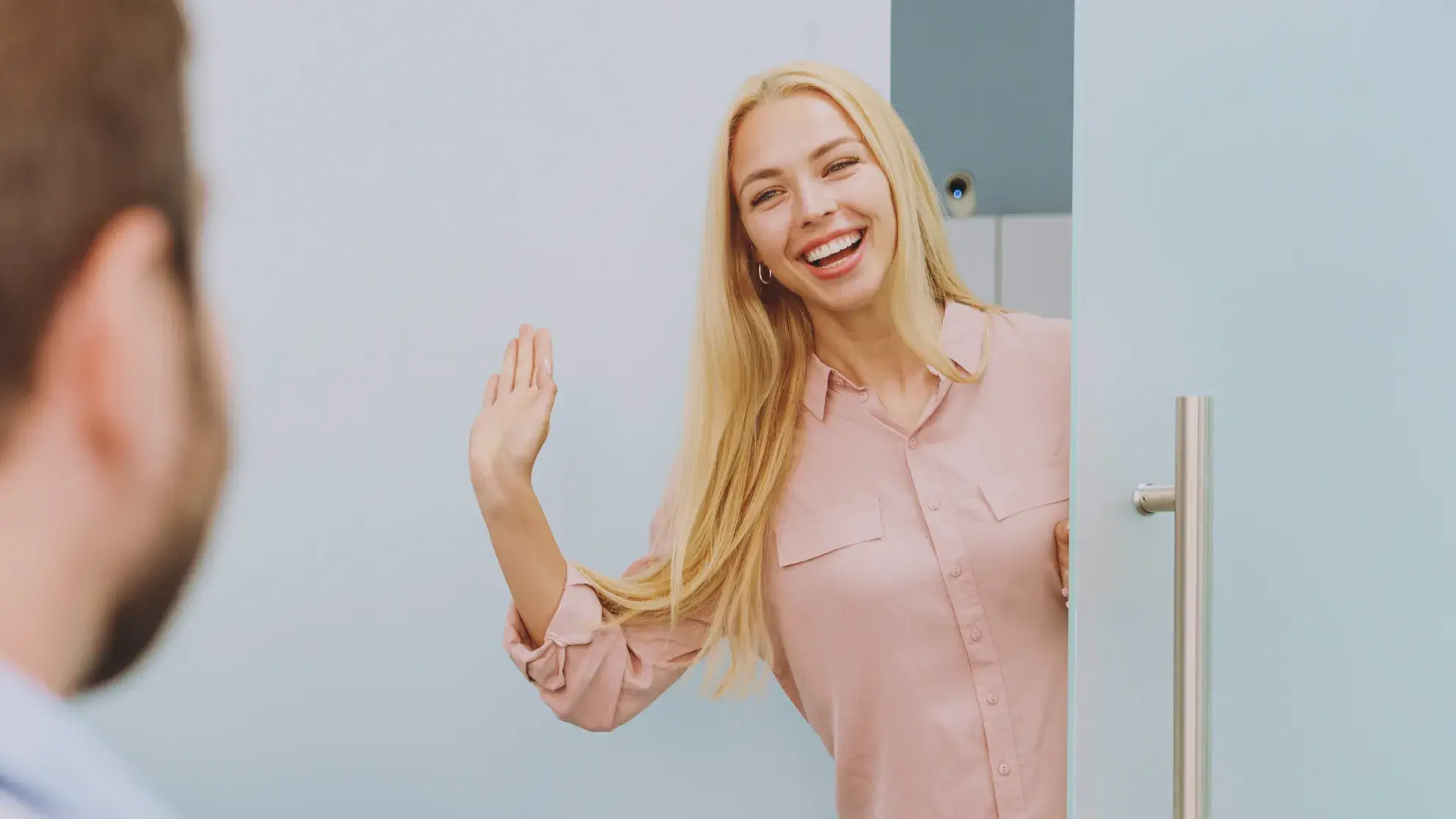 Smiling blonde woman waving while standing in a doorway greeting a man.