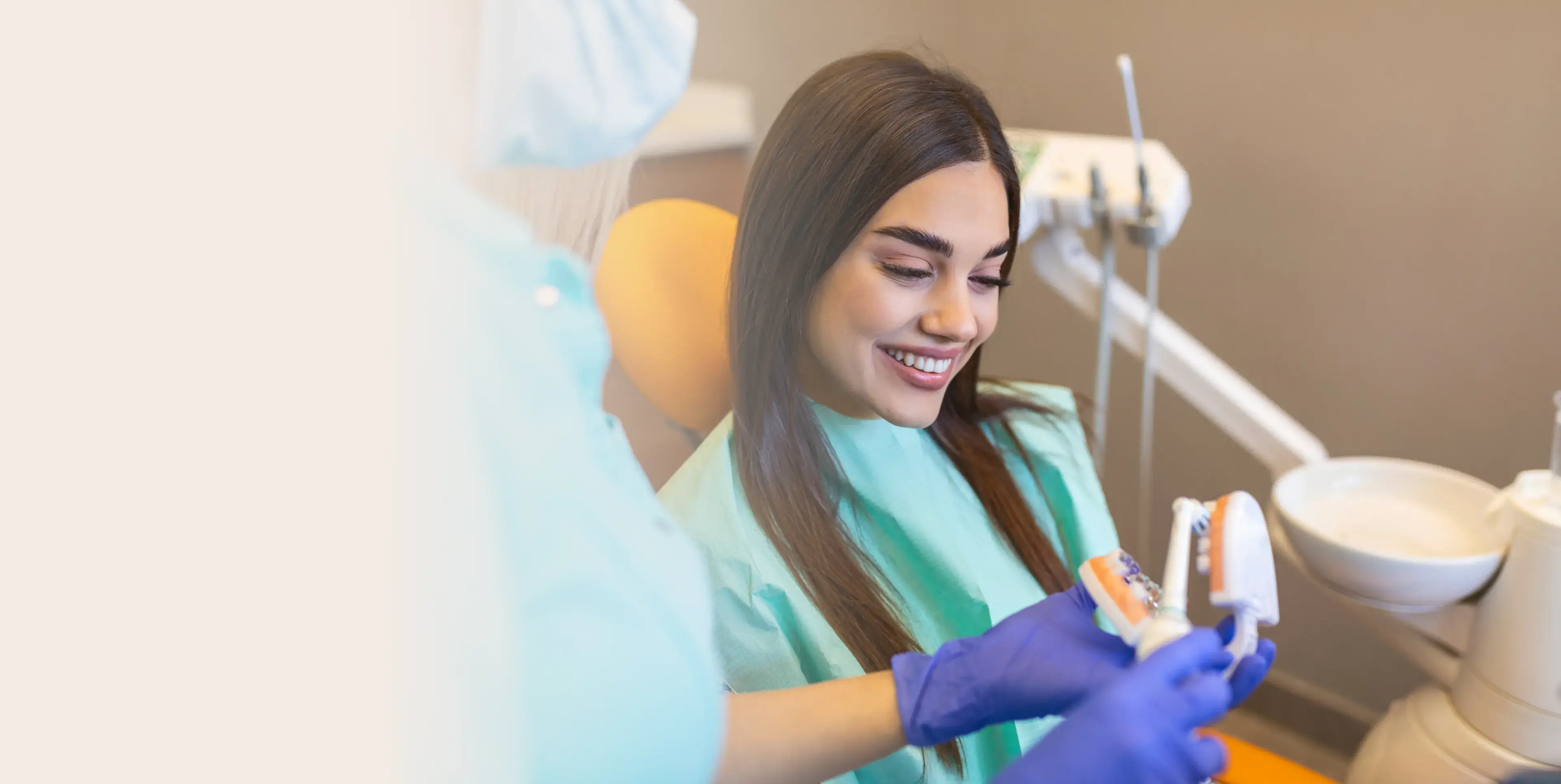 Smiling woman in a dental chair learning about oral hygiene from a dentist holding a tooth model and toothbrush.