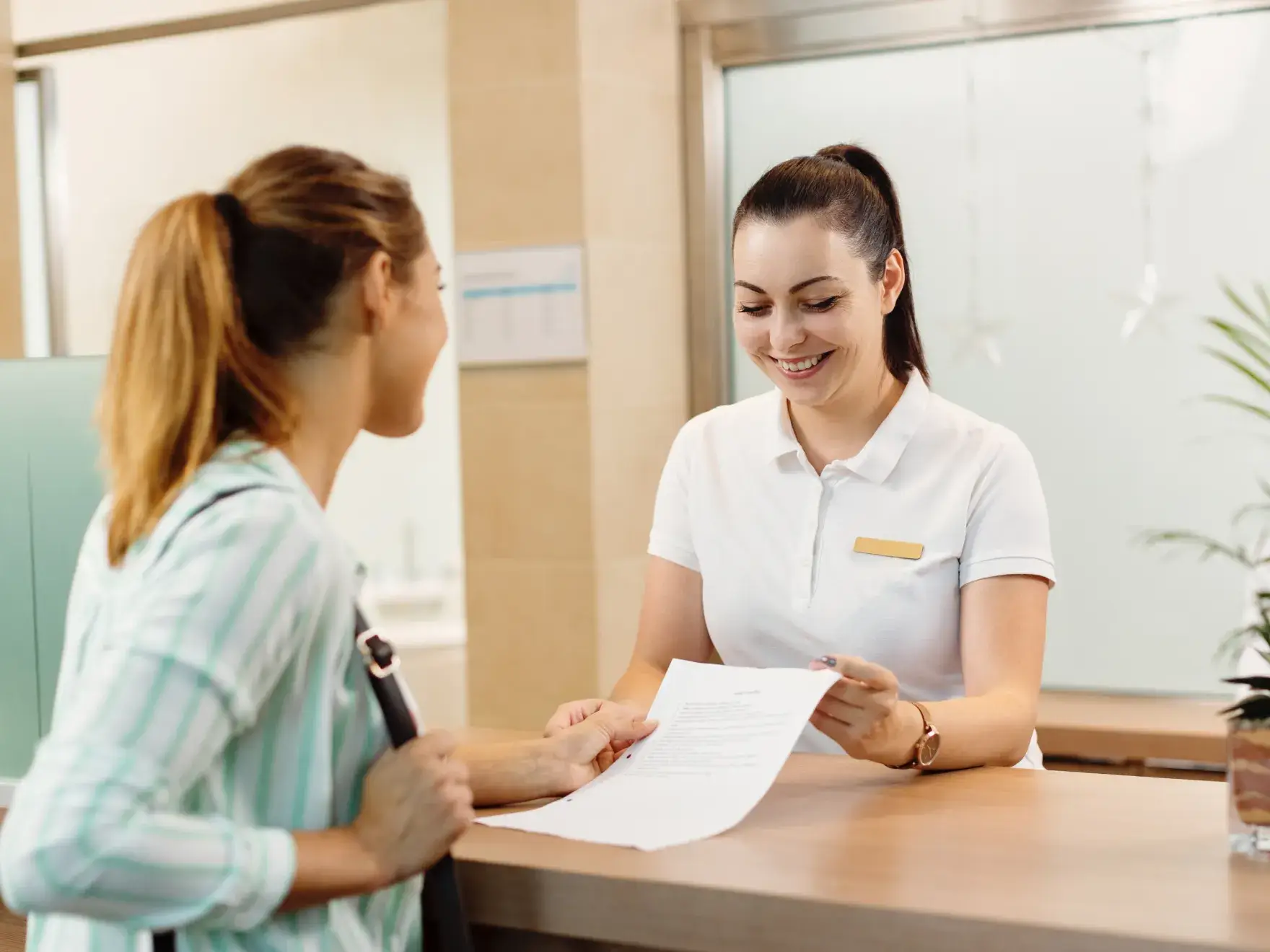 Smiling receptionist handing a document to a woman at a service desk.