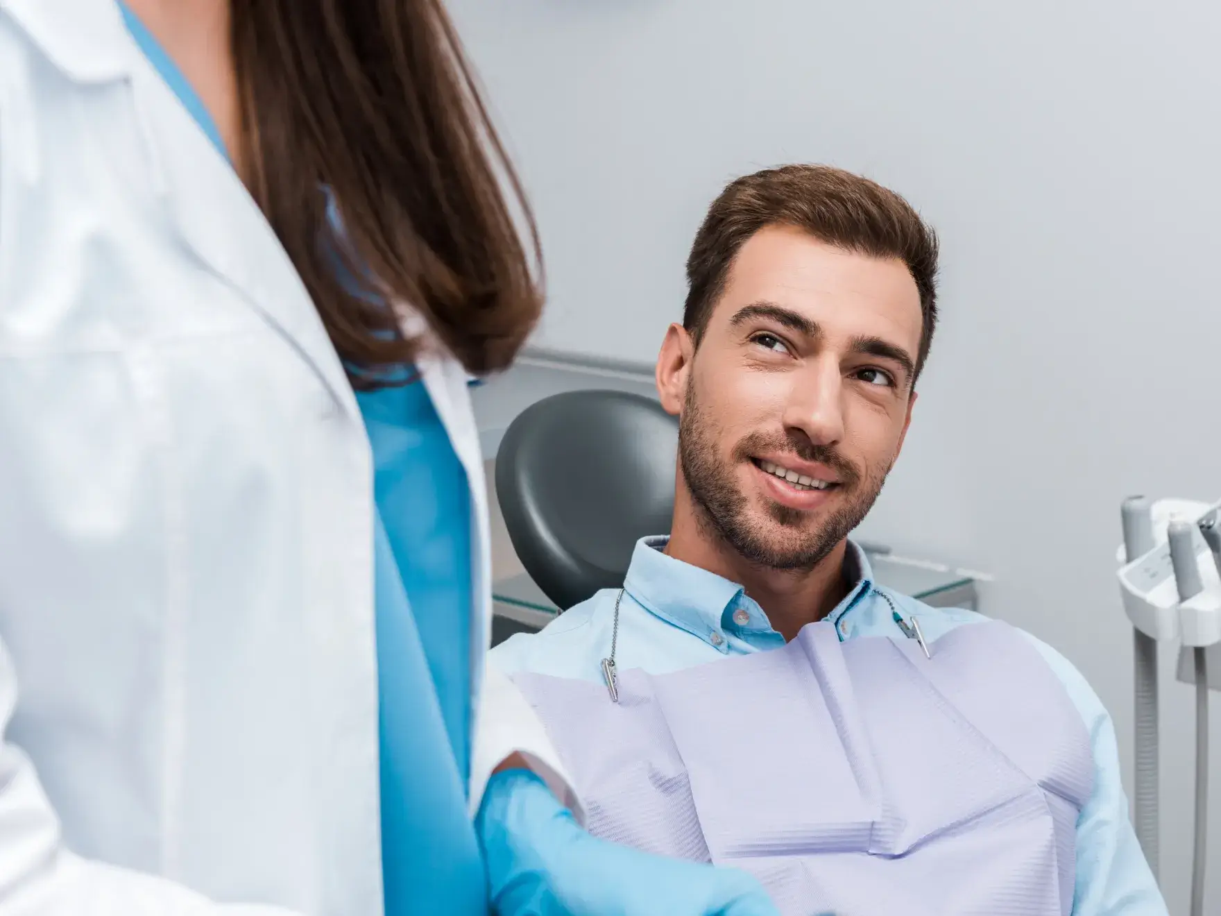 Smiling man sitting in dental chair looking at a female dentist wearing a white coat and blue gloves.