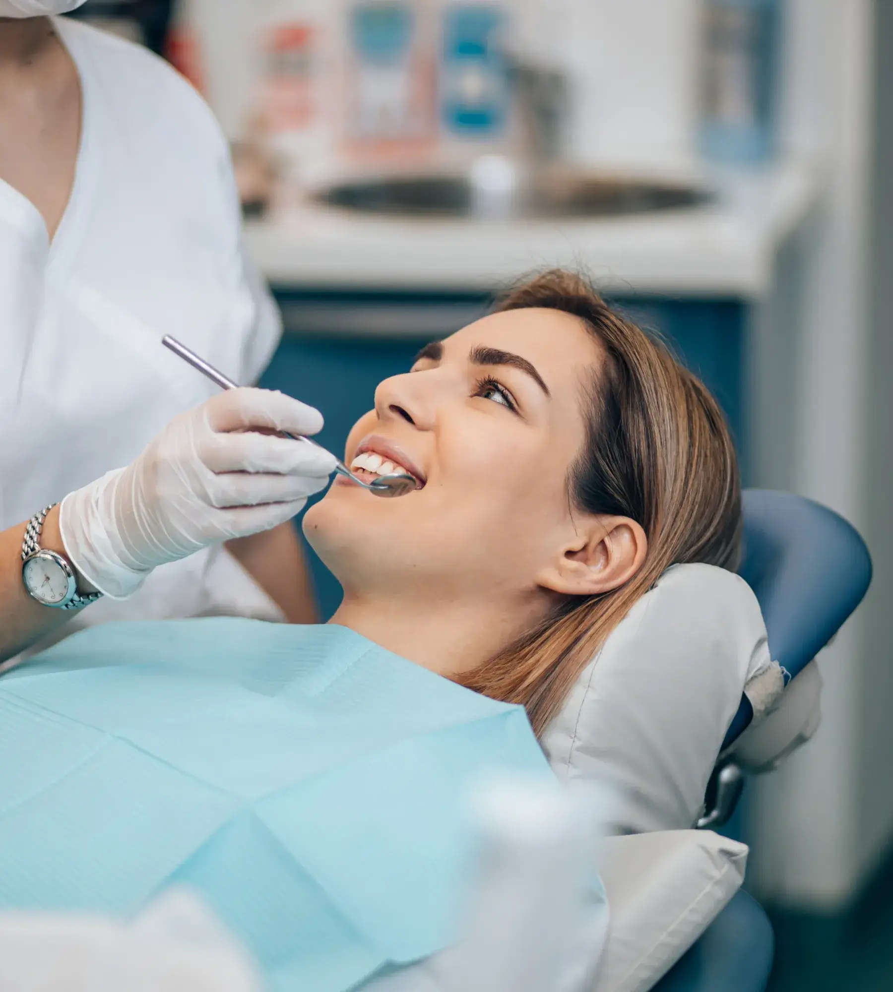 Young woman smiling in a dental chair with a dentist examining her teeth using a dental mirror.
