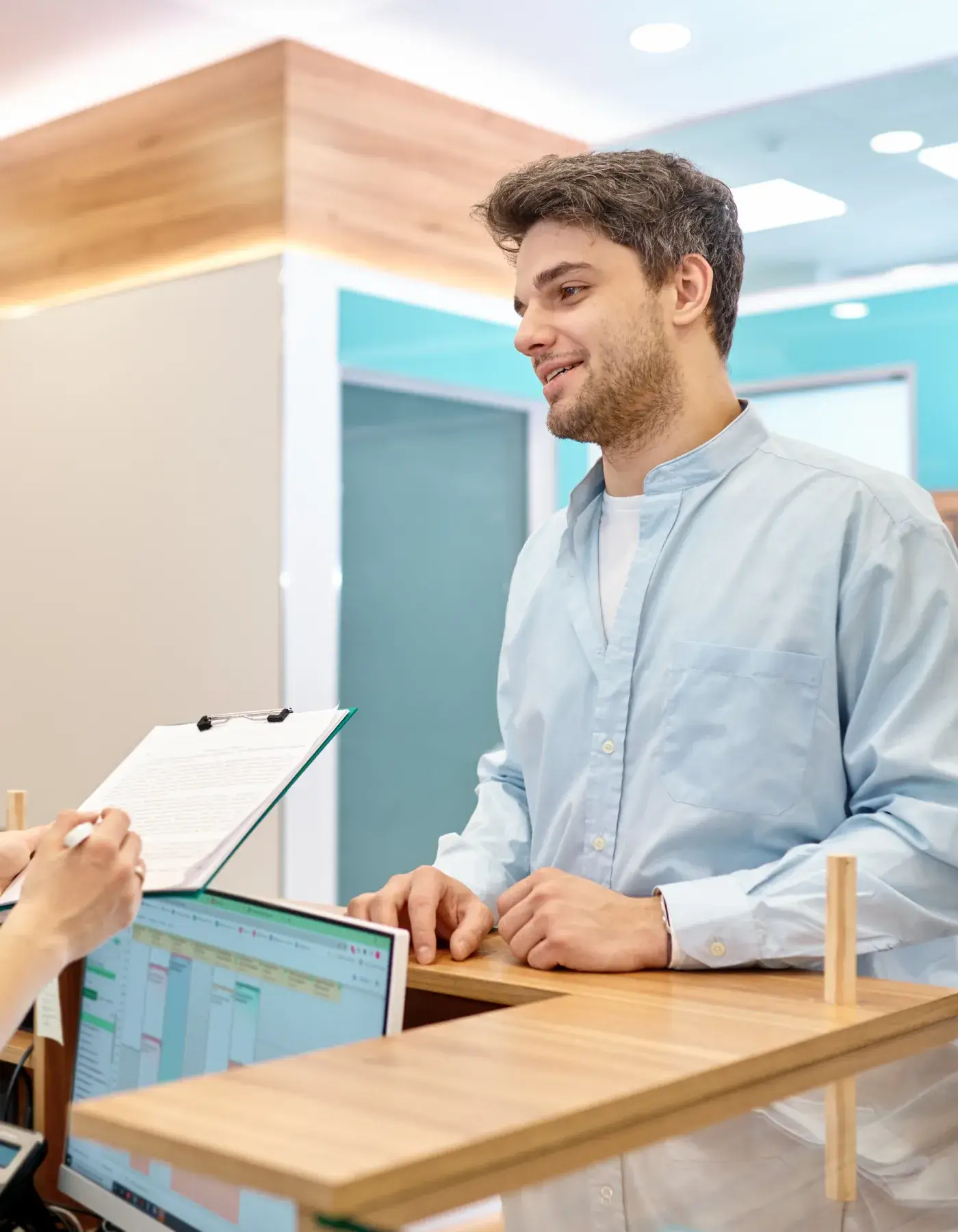 Young man in a light blue shirt standing at a reception desk, talking to a person holding a clipboard and pen.