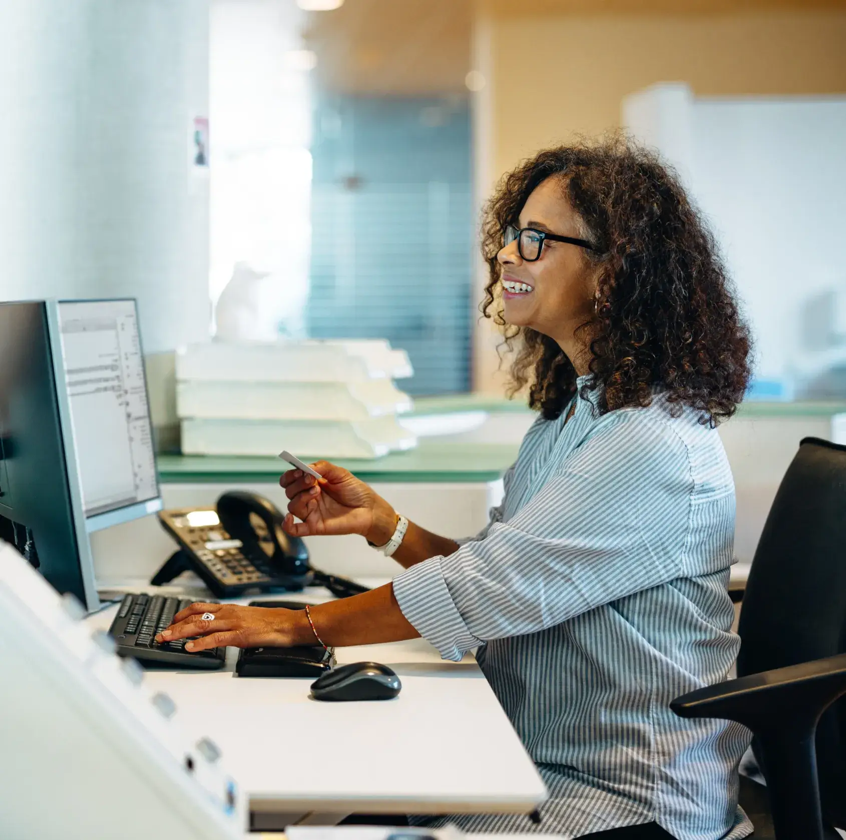 Smiling woman with curly hair and glasses working on a computer at an office desk.