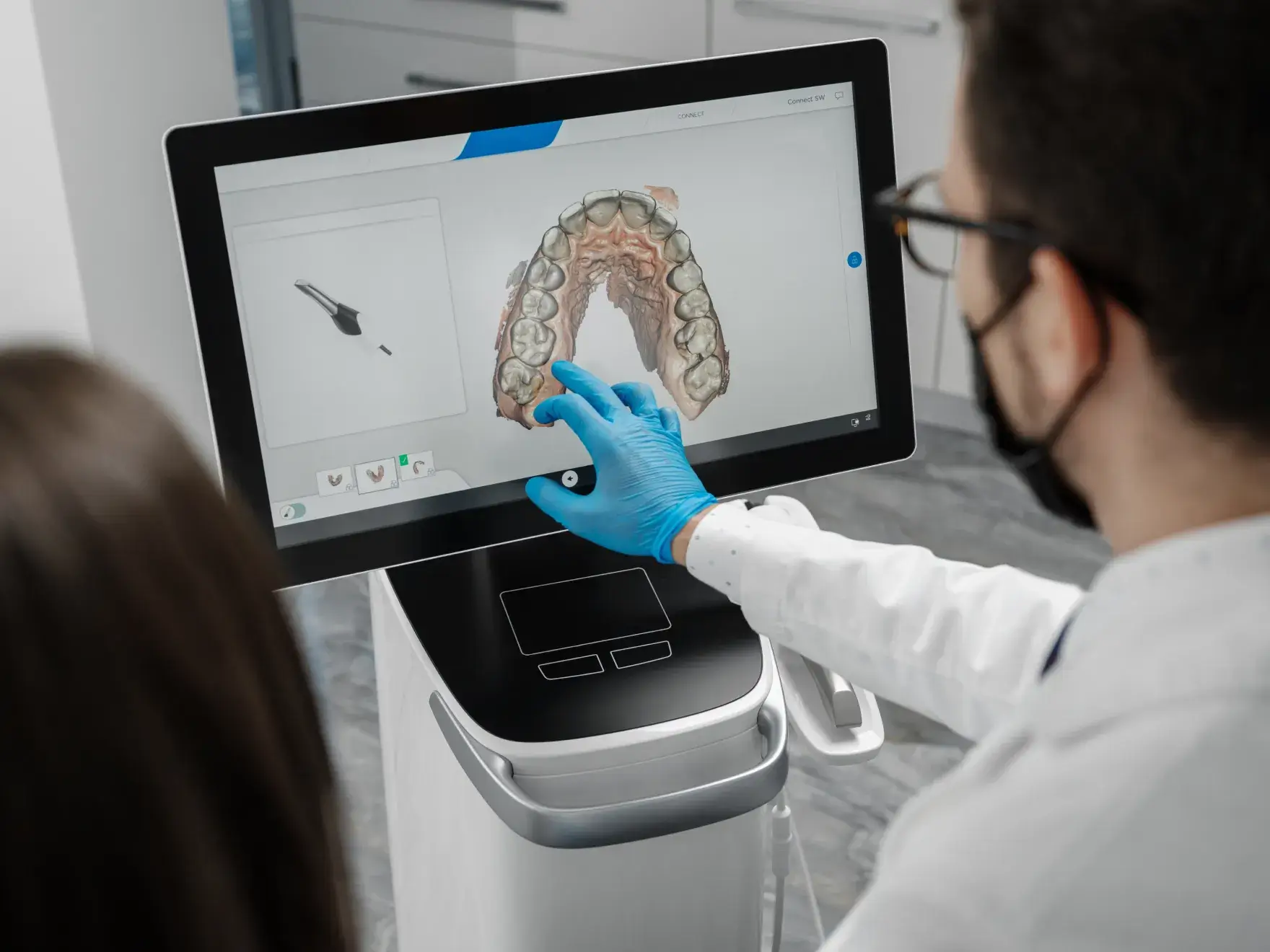 Dentist wearing blue gloves using a touchscreen to display a 3D digital model of upper teeth and gums.