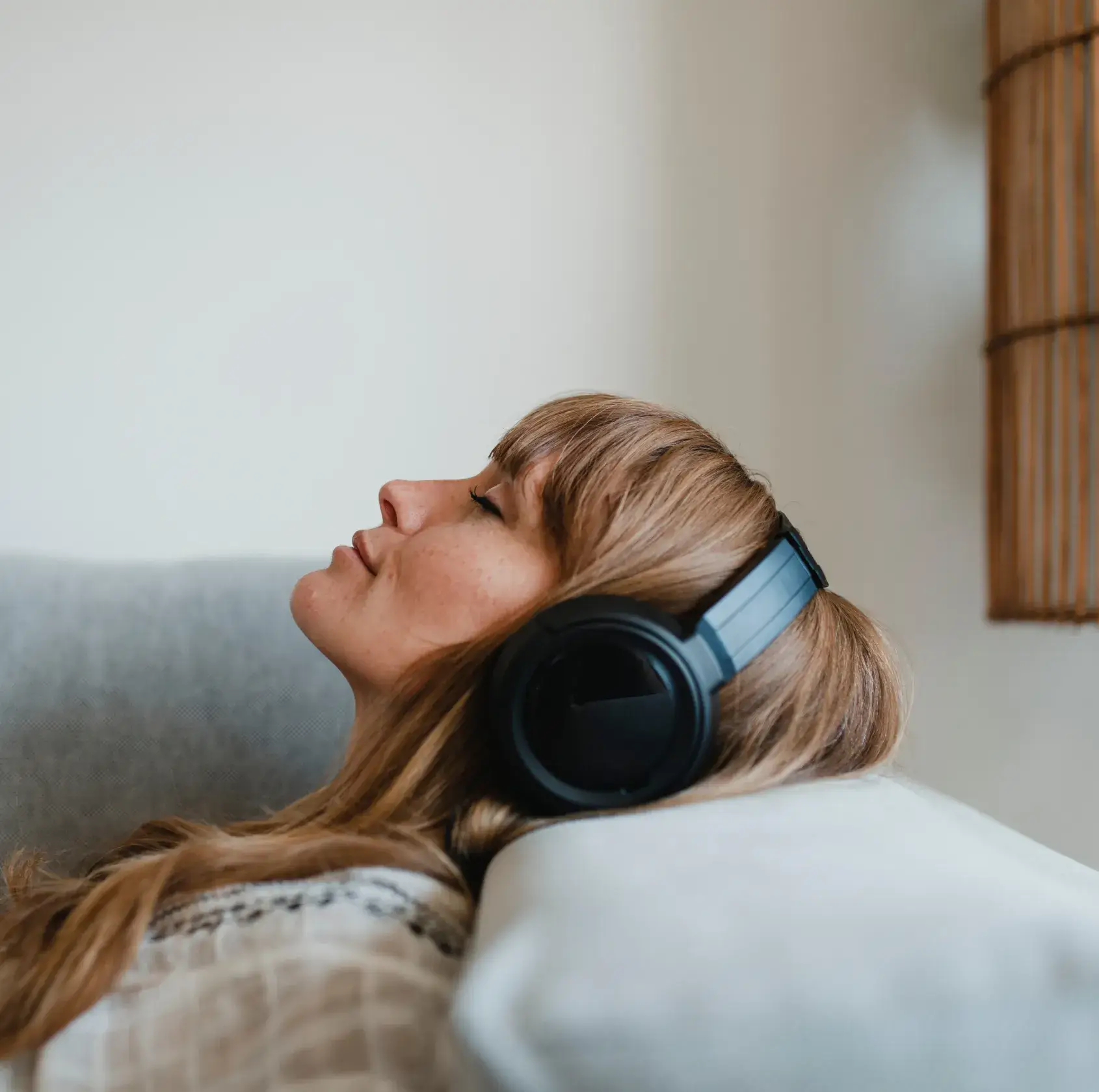 Relaxed woman with closed eyes listening to music on black headphones while lying on a light-colored sofa.