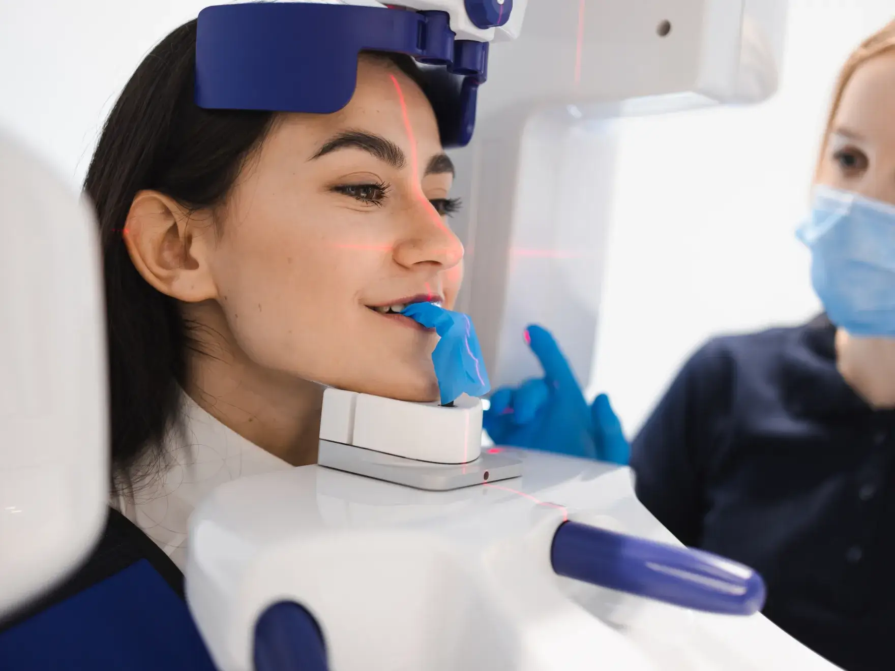 Woman undergoing a dental 3D scan with a blue bite impression in her mouth and a technician wearing a face mask in the background.