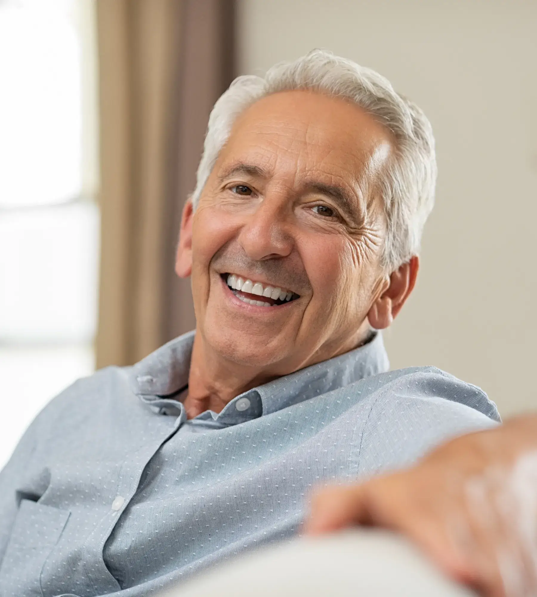 Smiling elderly man with gray hair wearing a light blue shirt, sitting comfortably.